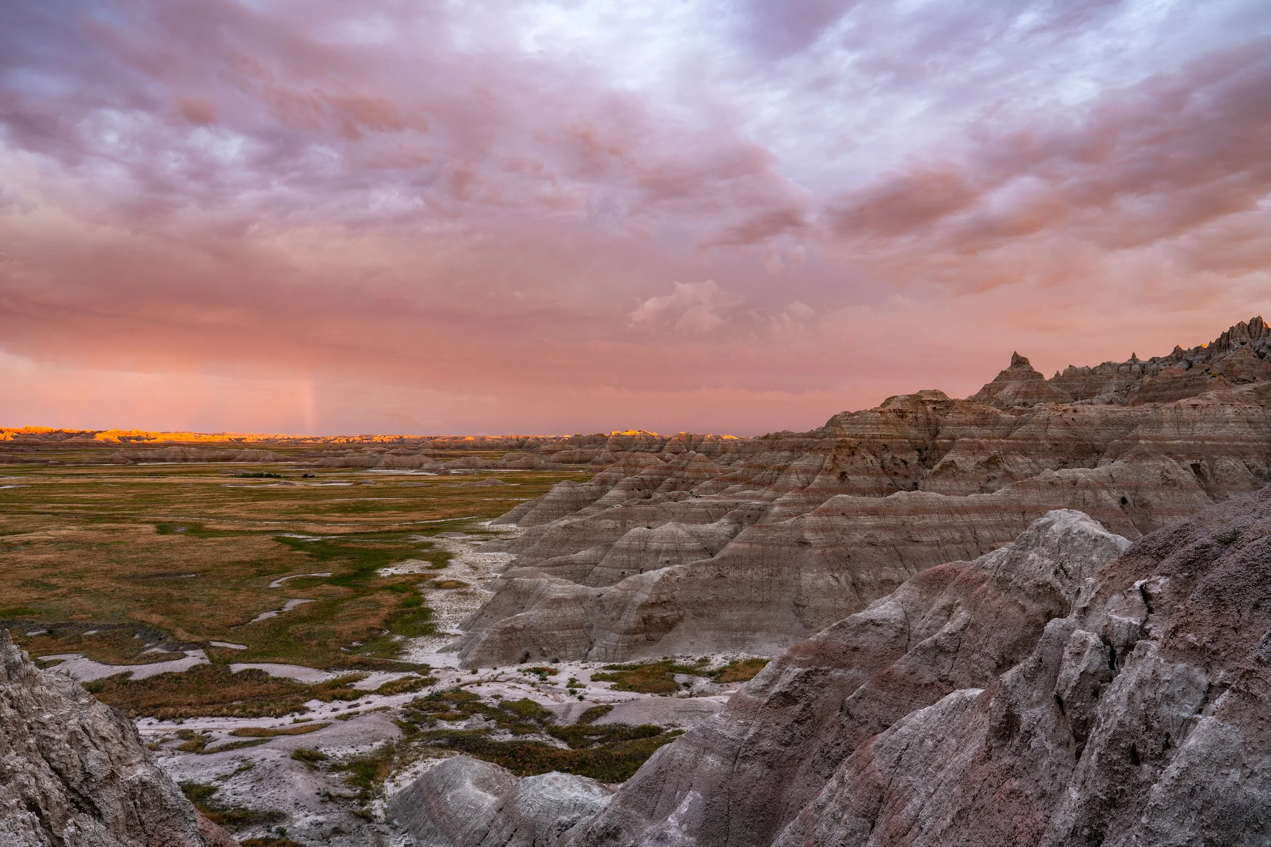 Landscape of badlands with layered rock formations, green patches, and a colorful sky with clouds at sunset or sunrise.