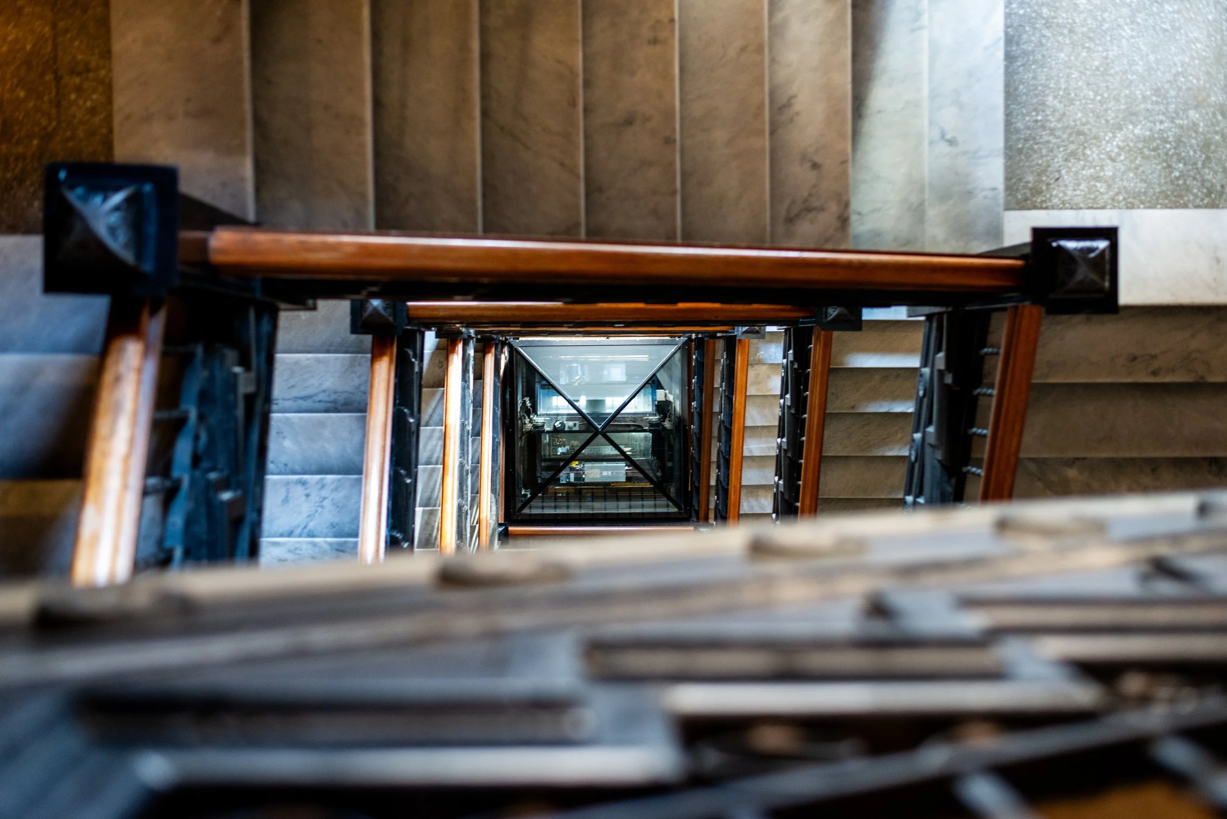 Looking down a spiral staircase from the top, showing the wooden handrails, black metal supports, and tiled walls on either side.