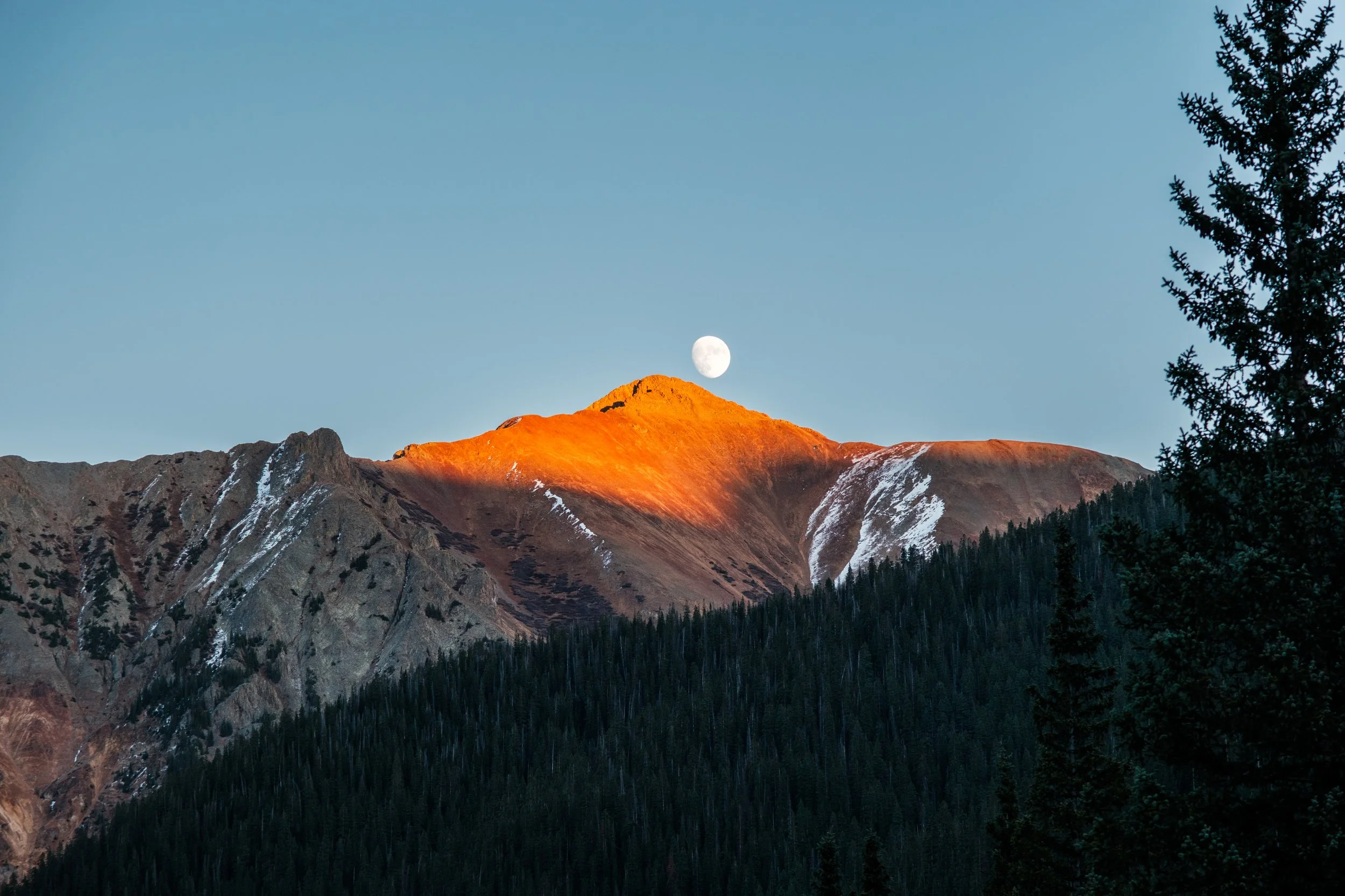 Mountain landscape at sunset with snow patches, dark forest at the base, a clear sky with a visible moon above the peak illuminated by orange sunlight.
