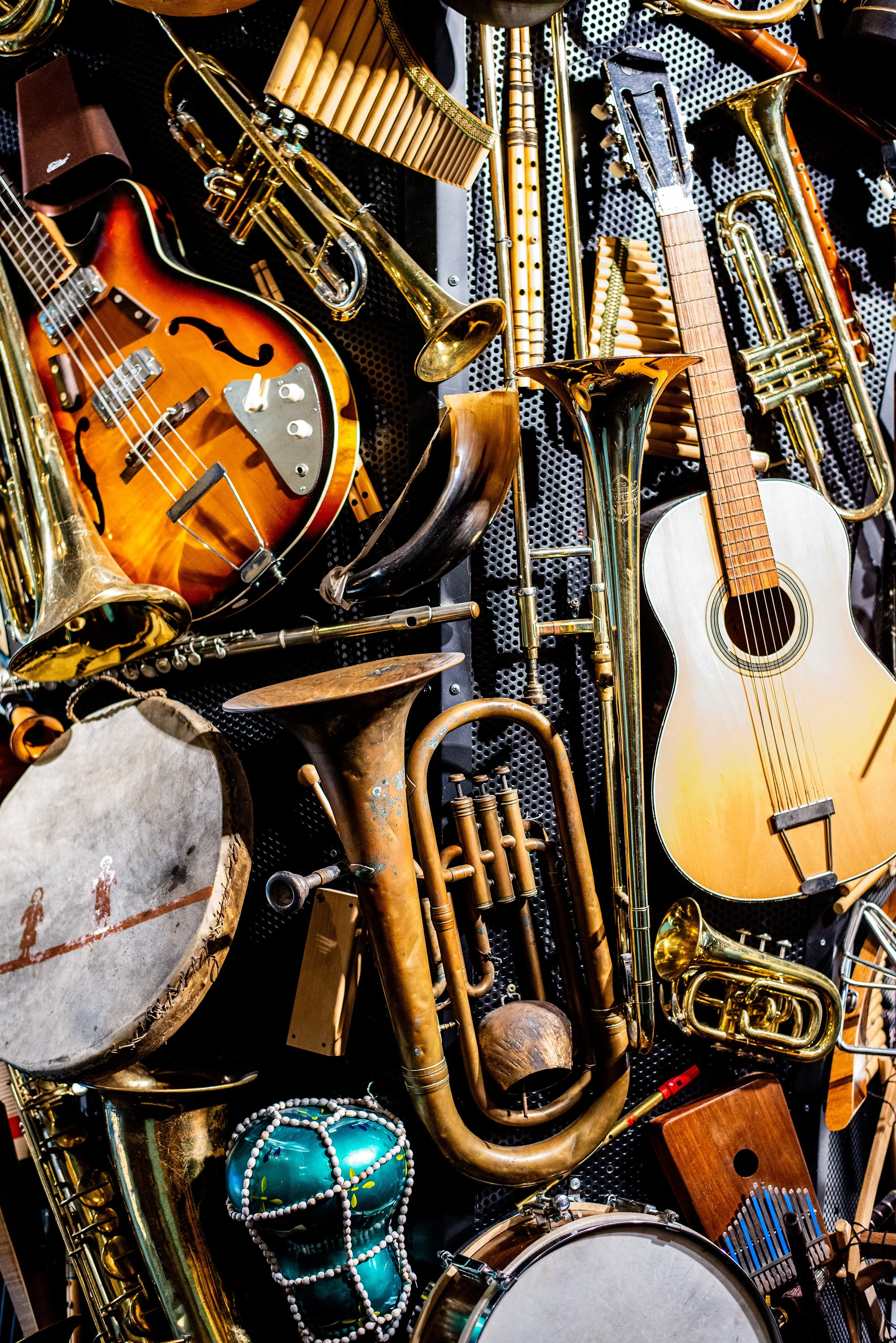 A collection of musical instruments including an acoustic guitar, saxophones, trumpets, trombones, a drum, a tambourine, and a maraca, arranged against a perforated black backdrop.