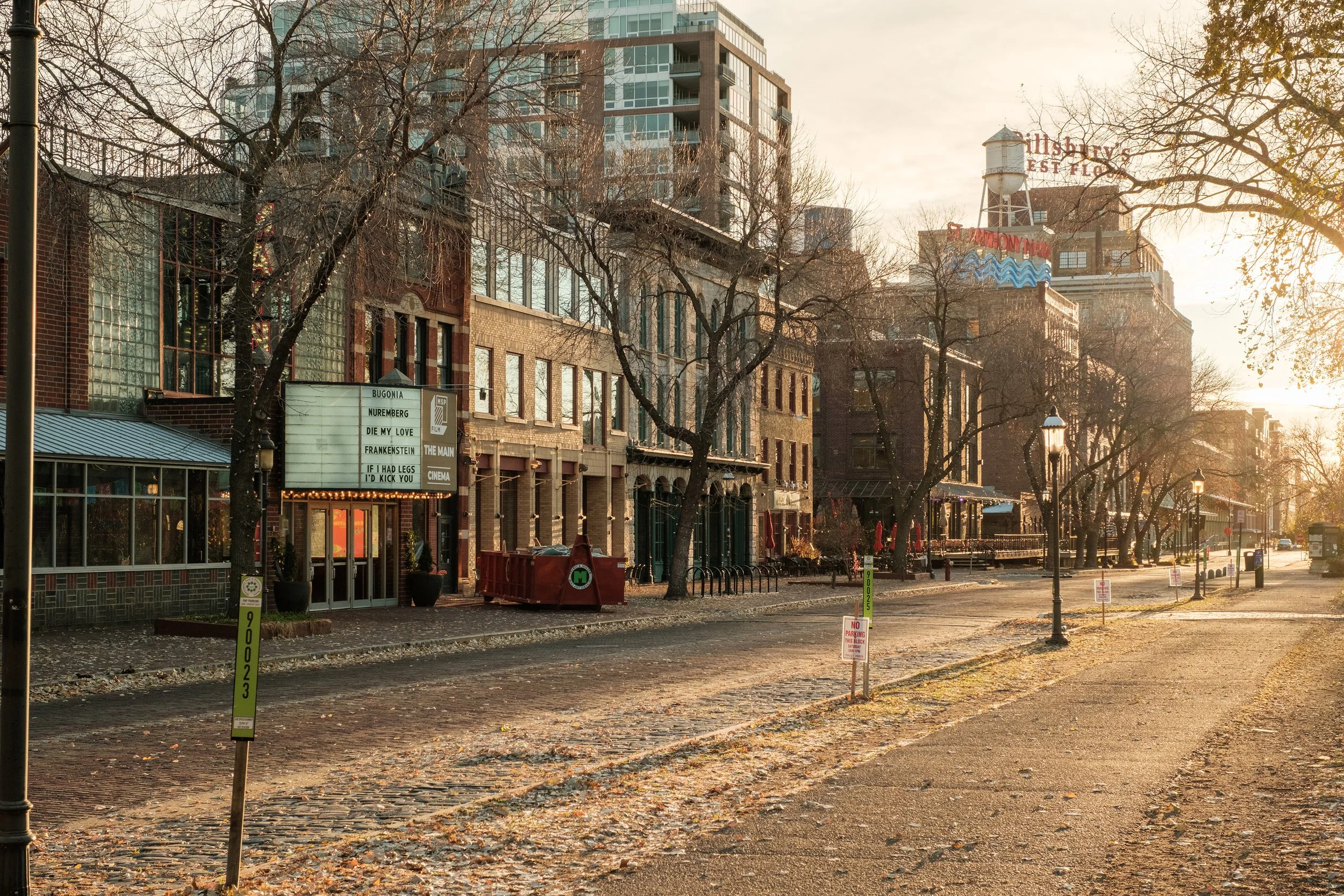 A quiet city street during sunset with bare trees, brick and modern buildings, street lamps, and parked bikes, illuminated by warm golden light.