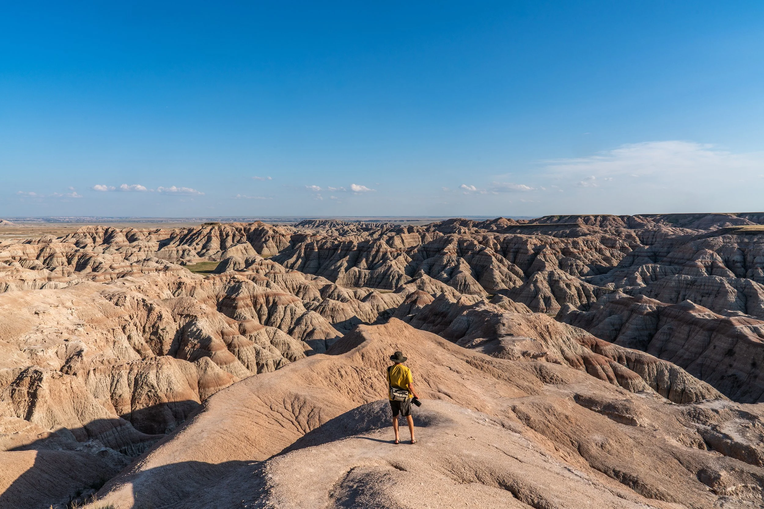 A person wearing a yellow jacket and a hat stands on a rock formation in a desert landscape with layered, eroded rock formations under a clear blue sky.