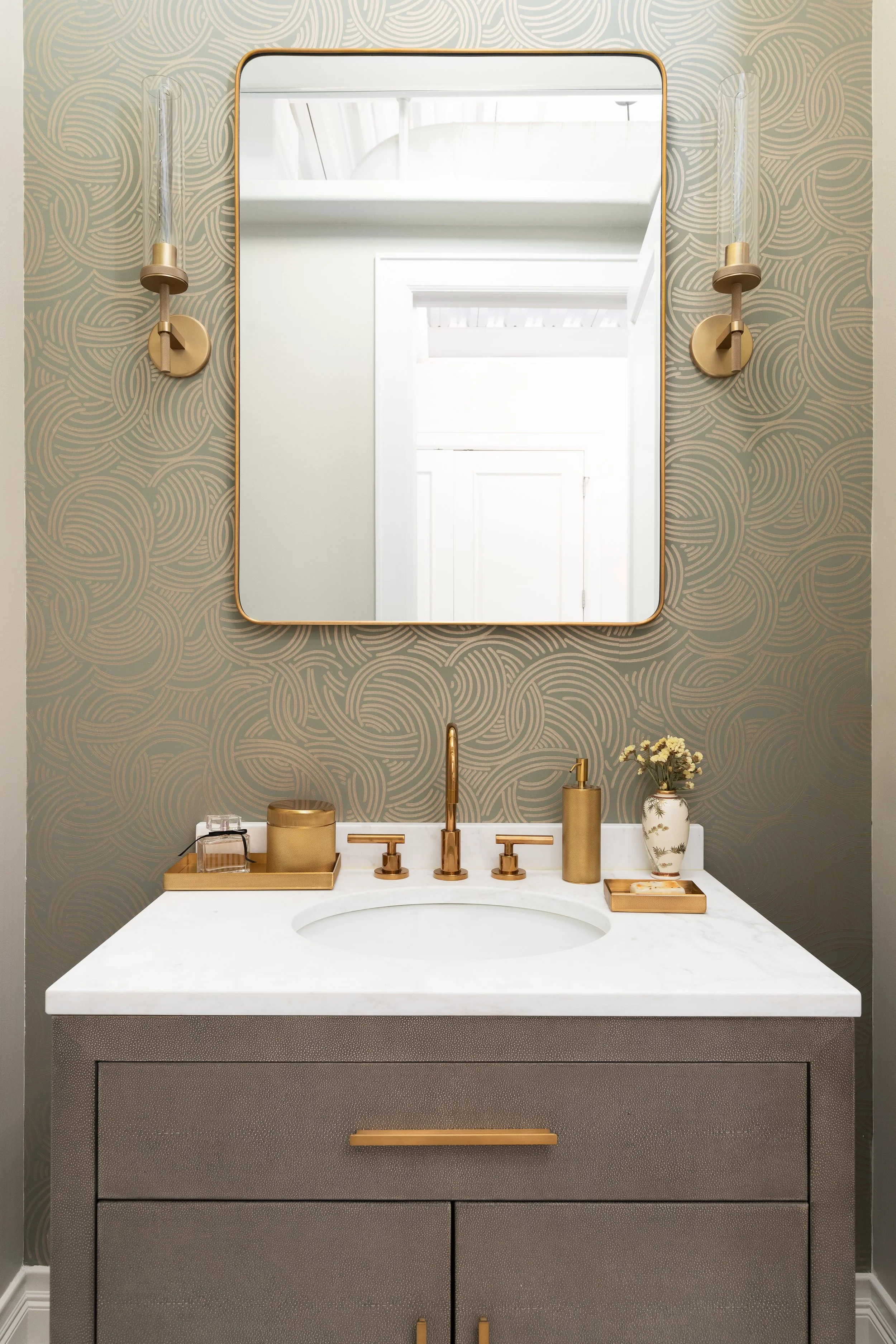 Bathroom vanity with a mirrored cabinet, gold fixtures, a marble countertop, and decorative toiletries, with patterned wallpaper in the background.