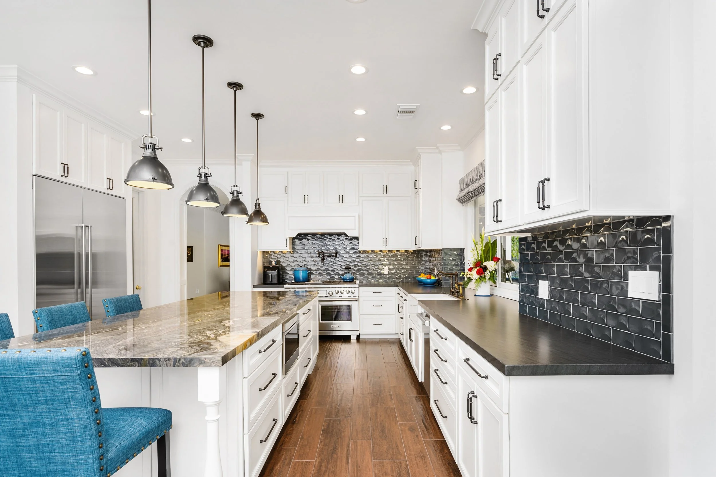 A modern kitchen with white cabinets, black handles, and a black tiled backsplash. There is a white countertop with a sink, a vase of flowers, and a blue bowl. The kitchen has wooden flooring and hanging pendant lights above a marble kitchen island with blue chairs.