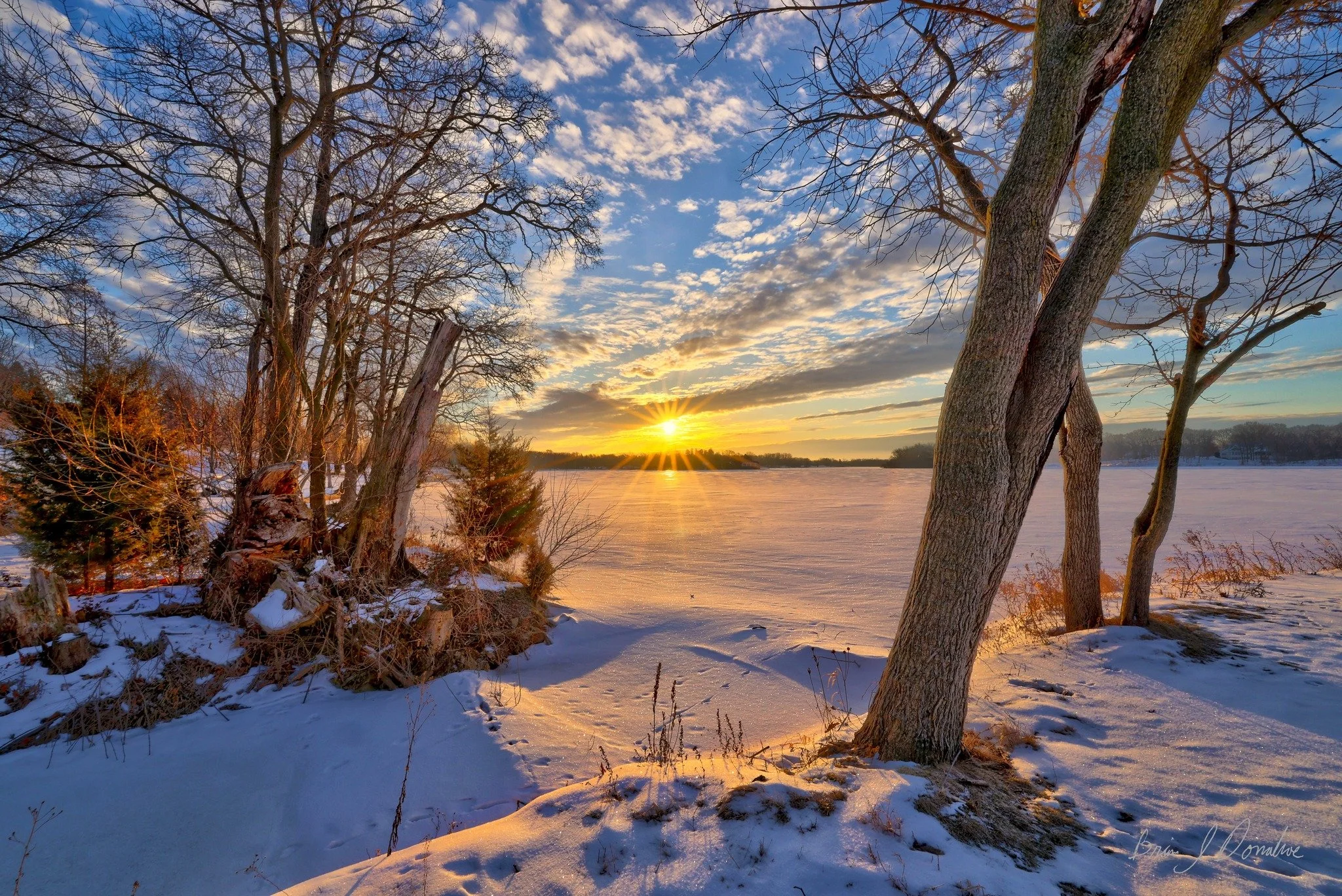 Snow-covered landscape with leafless trees by a frozen Countryside Lake at sunset, with clouds and a colorful sky.