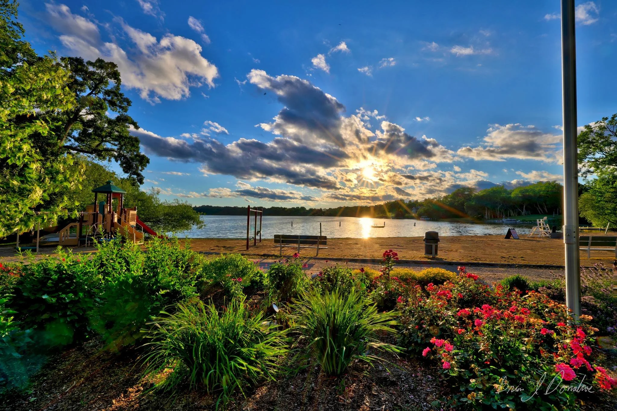 Sunset over Countryside Lake with a sandy beach, surrounded by green trees and a flower garden in the foreground.