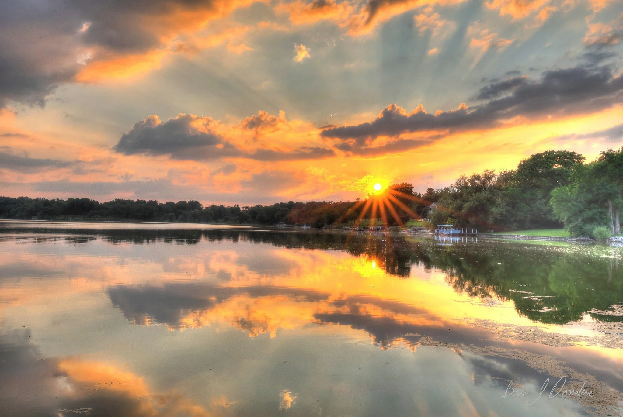 A peaceful lake sunset scene with the sun low on the horizon, casting a golden glow and starburst rays. The sky has orange, yellow, and pink clouds, reflected in the calm water. Trees line the shore on the right side.