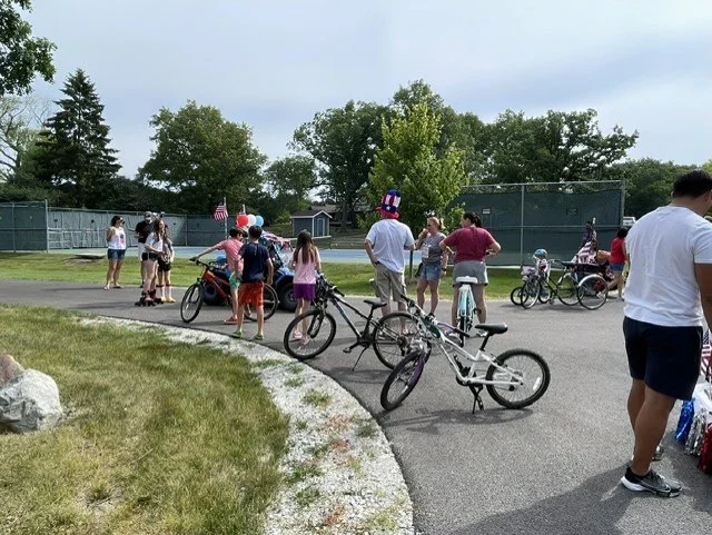 Countryside Lake Children and adults gathered in at the common area near the tennis court, some with bicycles, under a partly cloudy sky, with trees and a chain-link fence in the background.