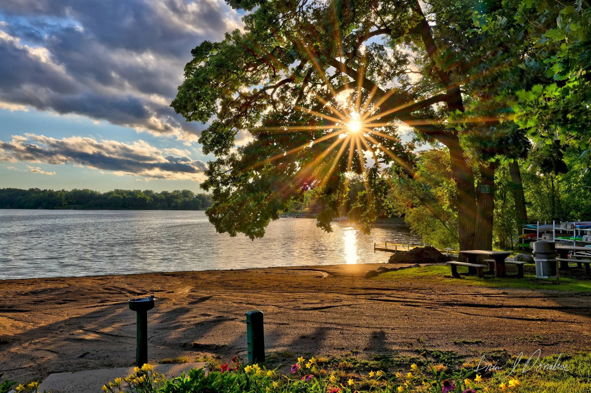 A scenic Countryside Lake view at sunset with the sun shining through the leaves of a large tree, casting rays of light. The lake is calm, lined with trees, and there are benches, a picnic table, and boats on the right side.