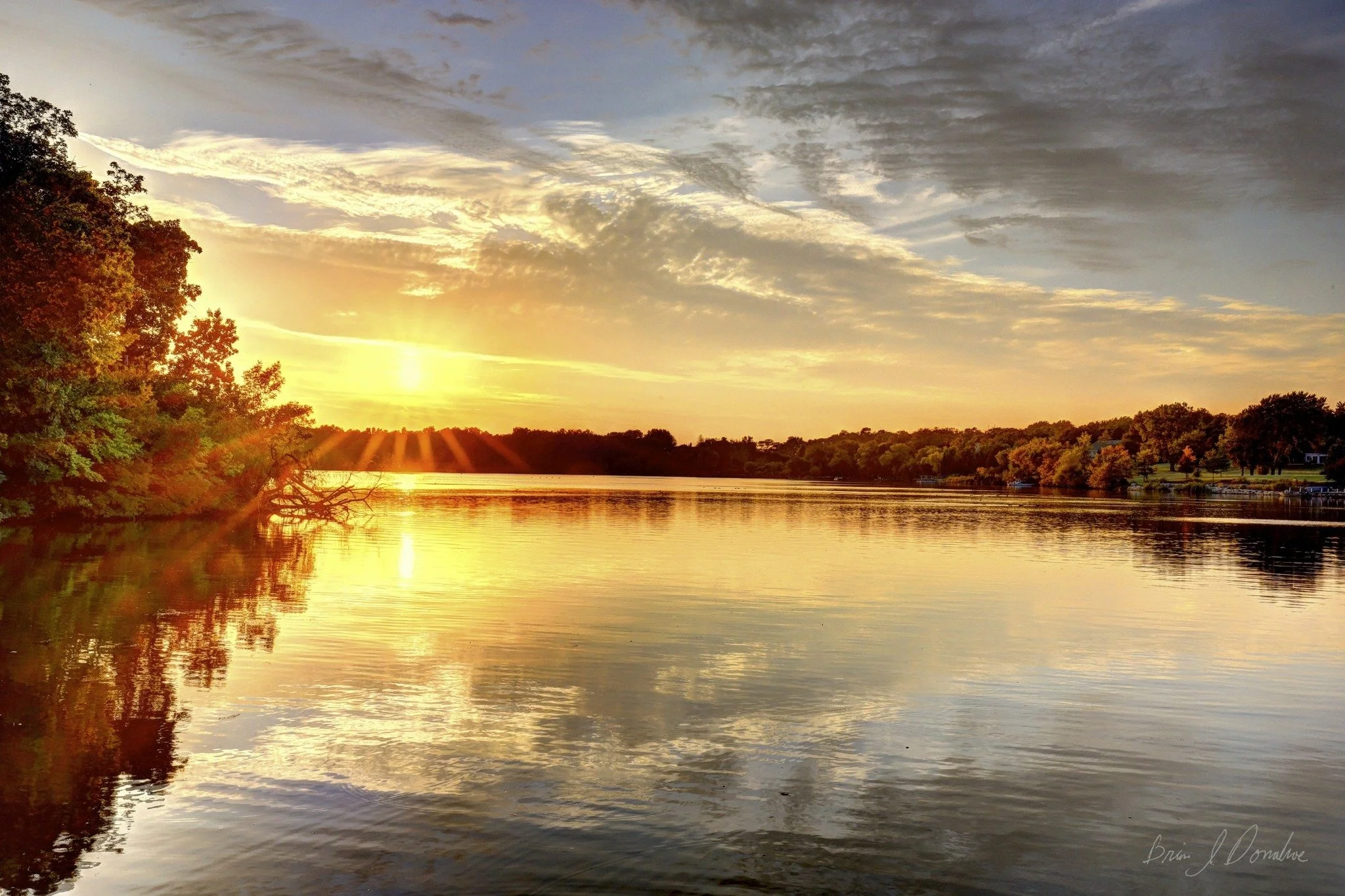 Serene Countryside Lake at sunset with cloudy sky, trees along the shore reflecting in the water, and sun rays illuminating the scene.
