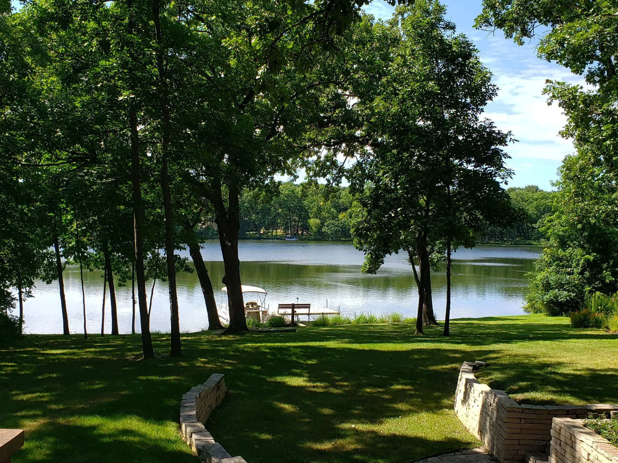 View of a Countryside Lake from a resident's back yard with trees surrounding it, a bench, and a boat docked near the shore on a sunny day.