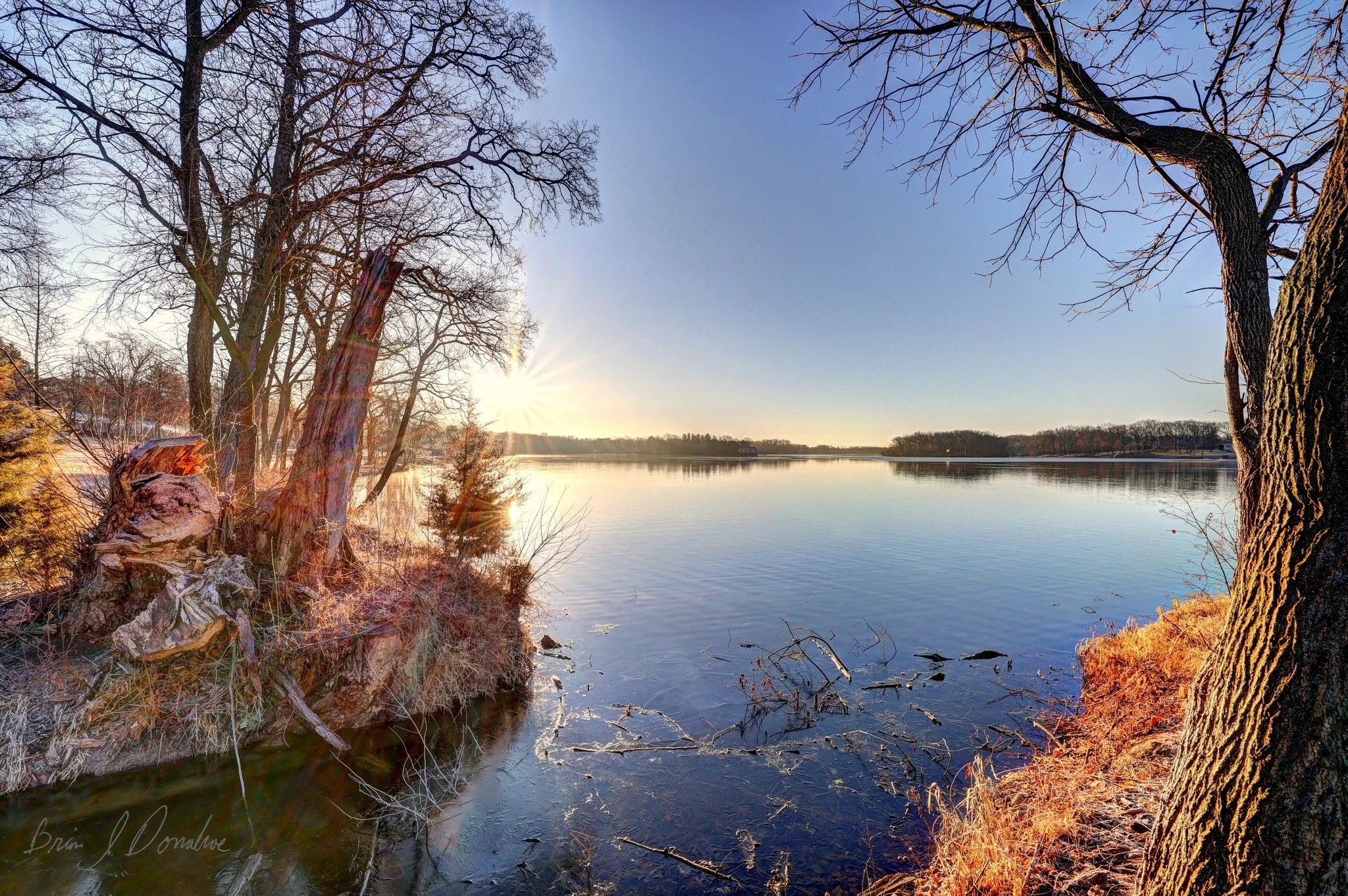 A peaceful lakeside scene during sunset with bare trees along the shore and calm water reflecting the sky.