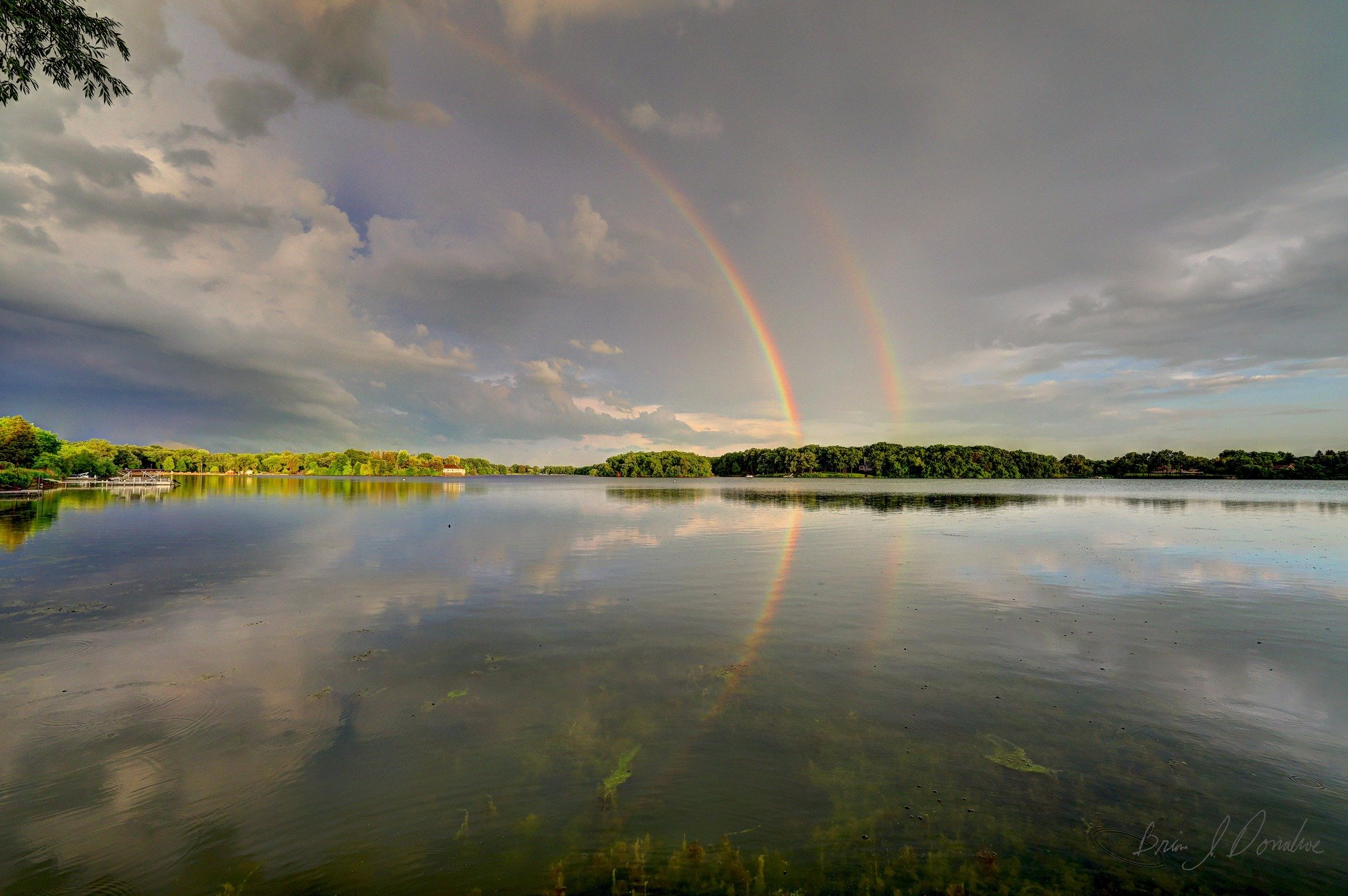 Countryside Lake with a rainbow in the sky, reflecting on the water, with trees along the shoreline and cloudy weather.