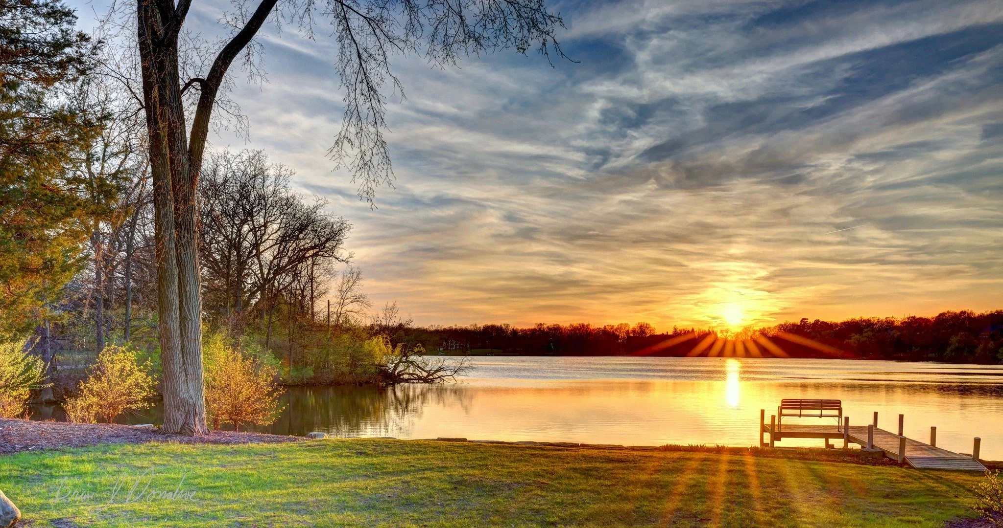 Sunset over Countryside Lake with a wooden dock, surrounded by trees and grassy area in the foreground.