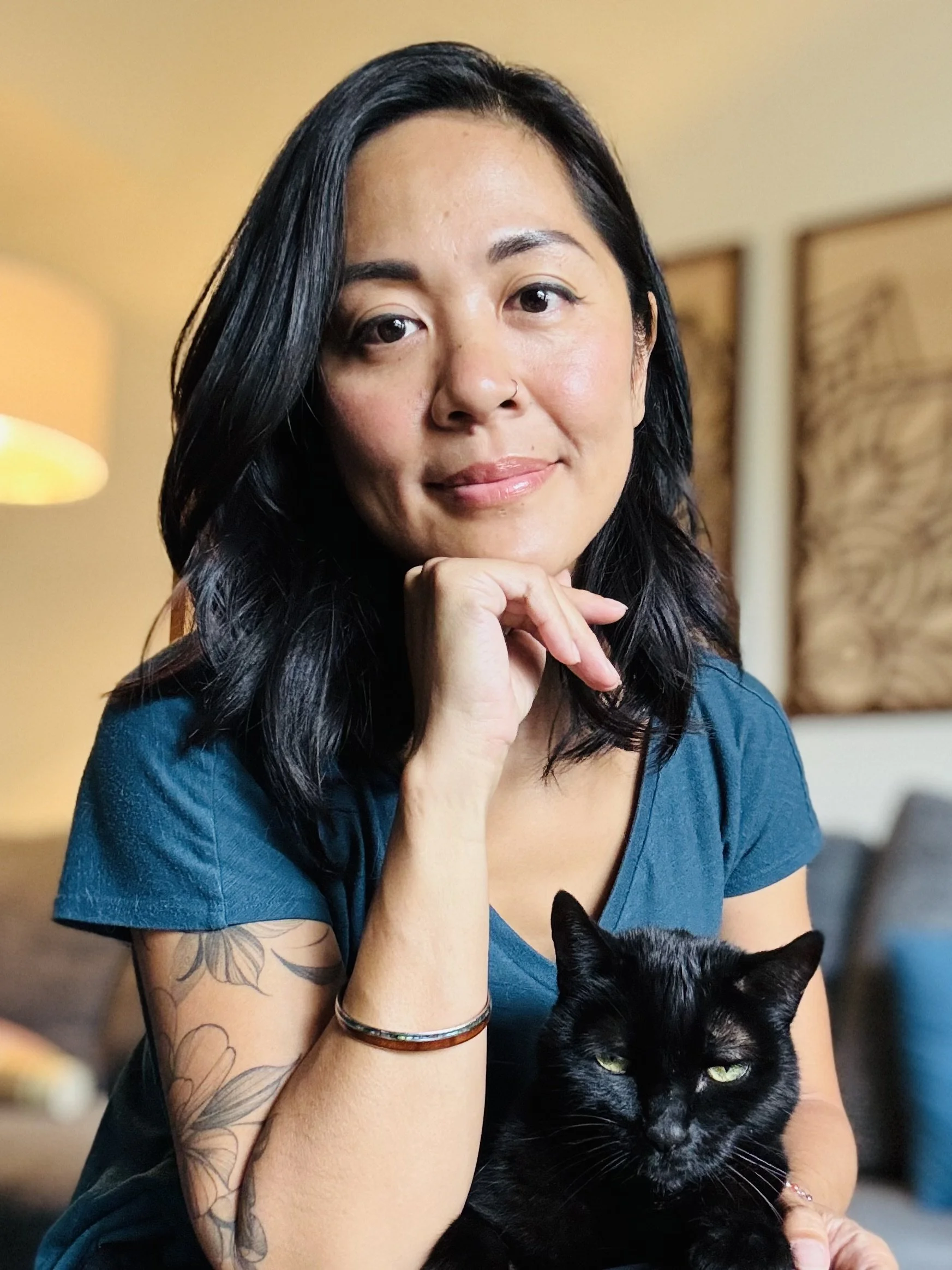 A woman with long black hair, wearing a blue shirt, sitting in a cozy indoor space with artwork on the wall behind her, holding a black cat with bright green eyes.