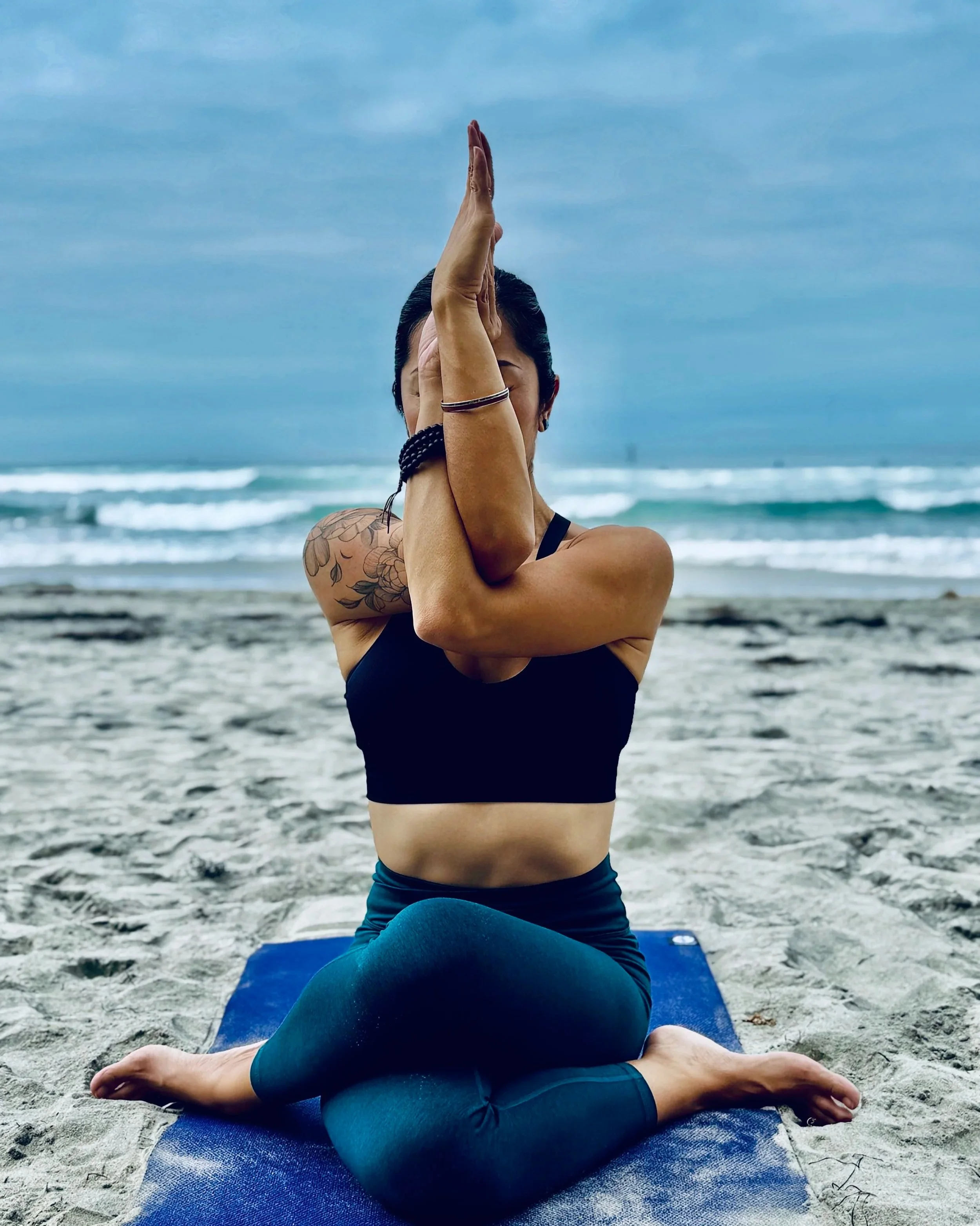 A person practicing yoga on a blue mat on the beach, sitting in a cross-legged position with hands in prayer pose overhead, facing the ocean with waves and a cloudy sky in the background.