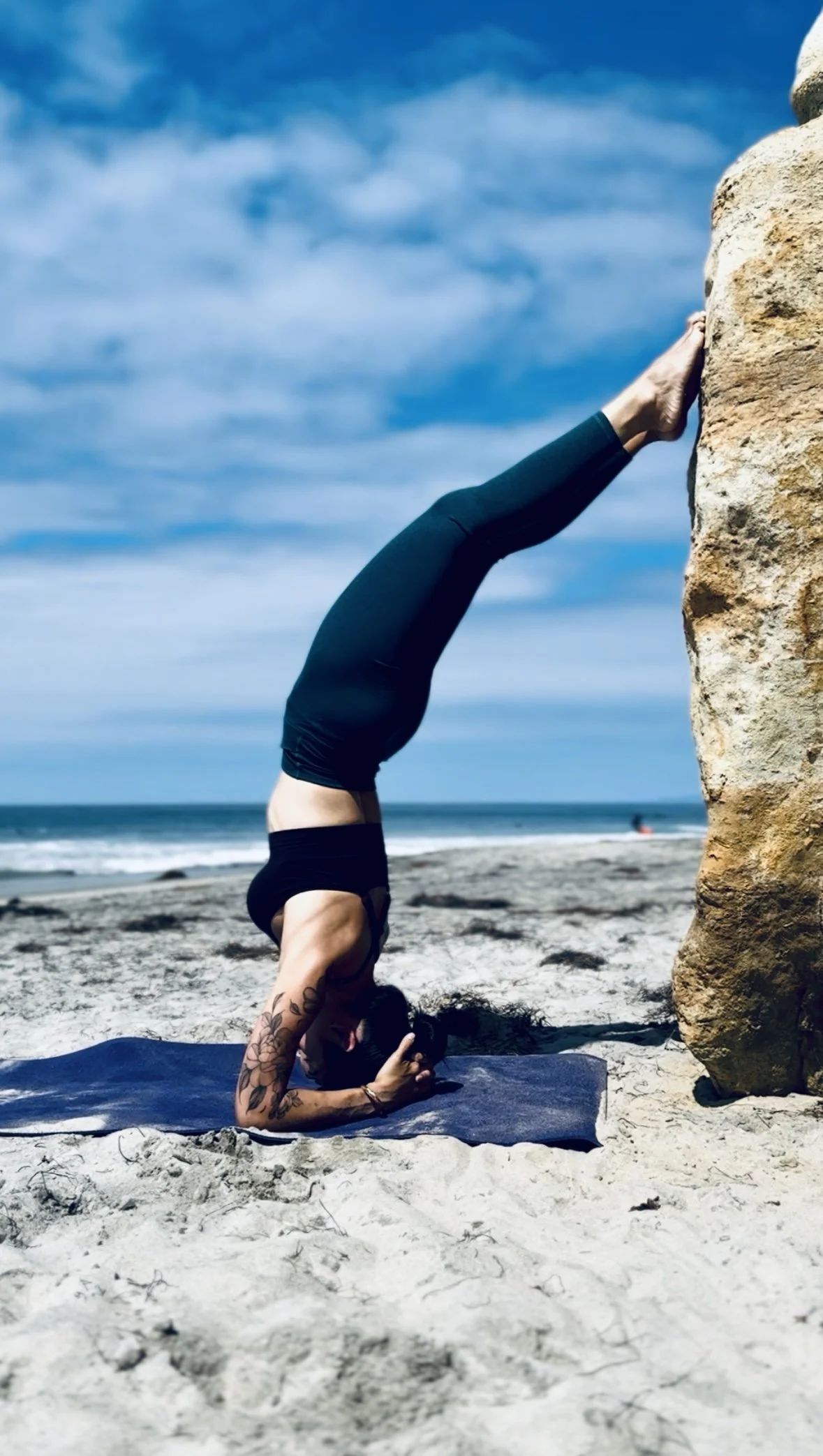 A woman doing a headstand yoga pose on a beach, with her back arched over her head, her feet against a large rock, and ocean waves and a blue sky in the background.