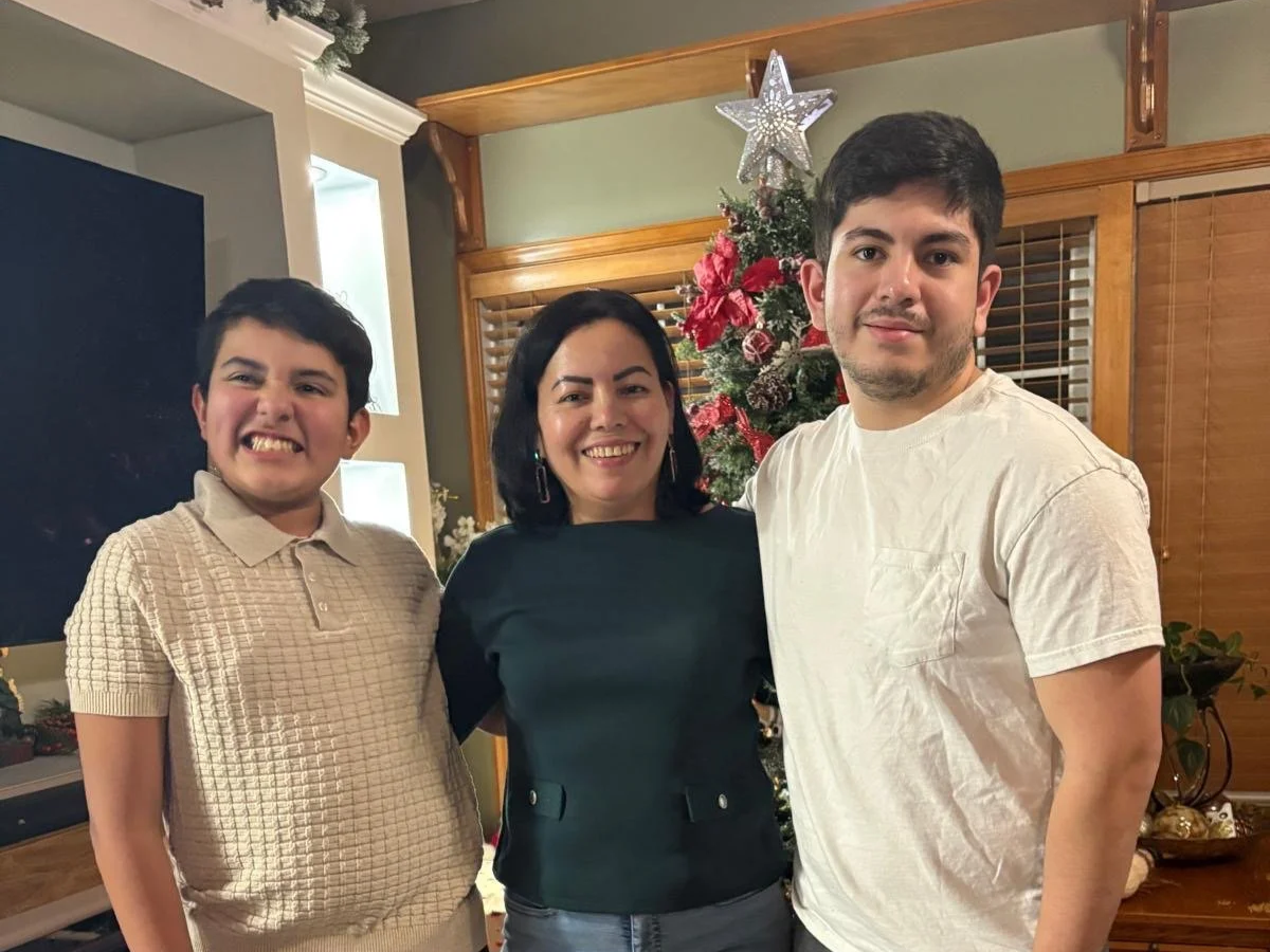 A woman and two young men standing in front of a decorated Christmas tree in a living room, smiling at the camera.
