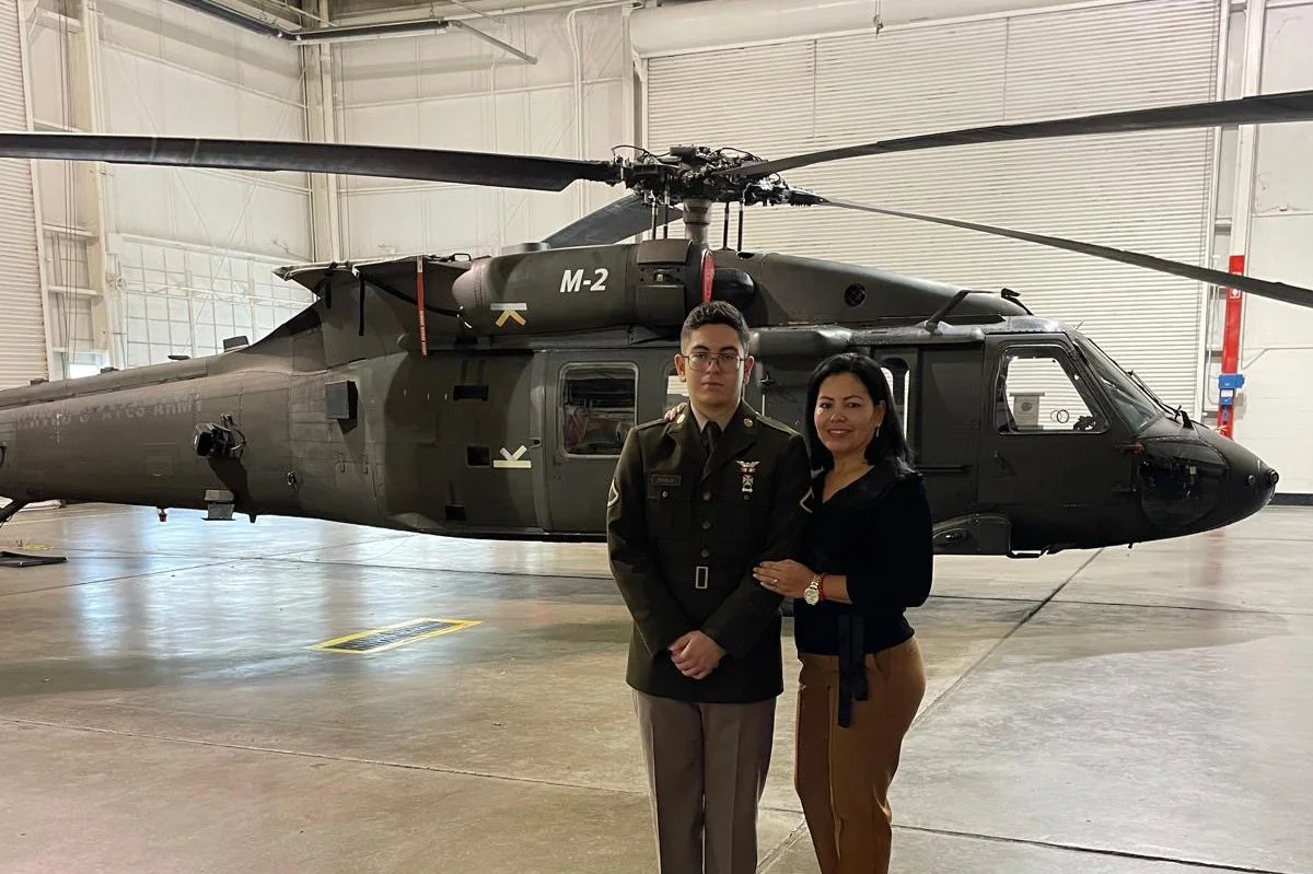 A man in a military uniform and a woman in casual clothing posing in front of a military helicopter inside a hangar.