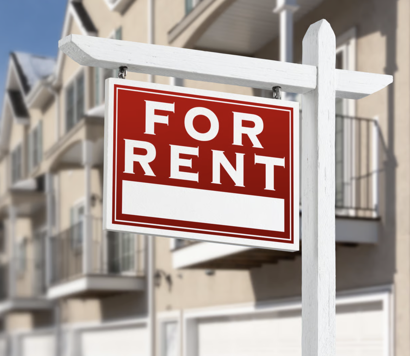 A 'For Rent' sign hanging on a white wooden post in front of a residential building.