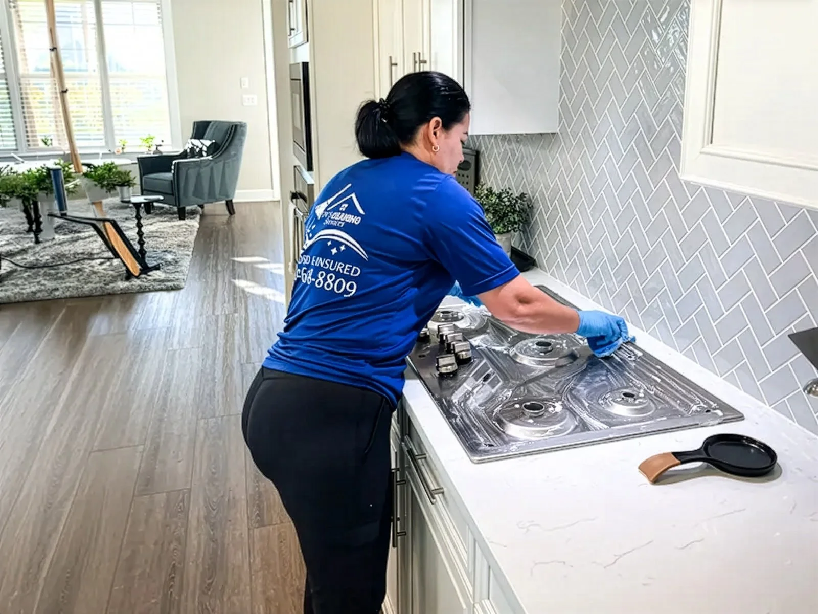 A woman wearing a blue shirt and black pants cleaning a stove in a modern kitchen.
