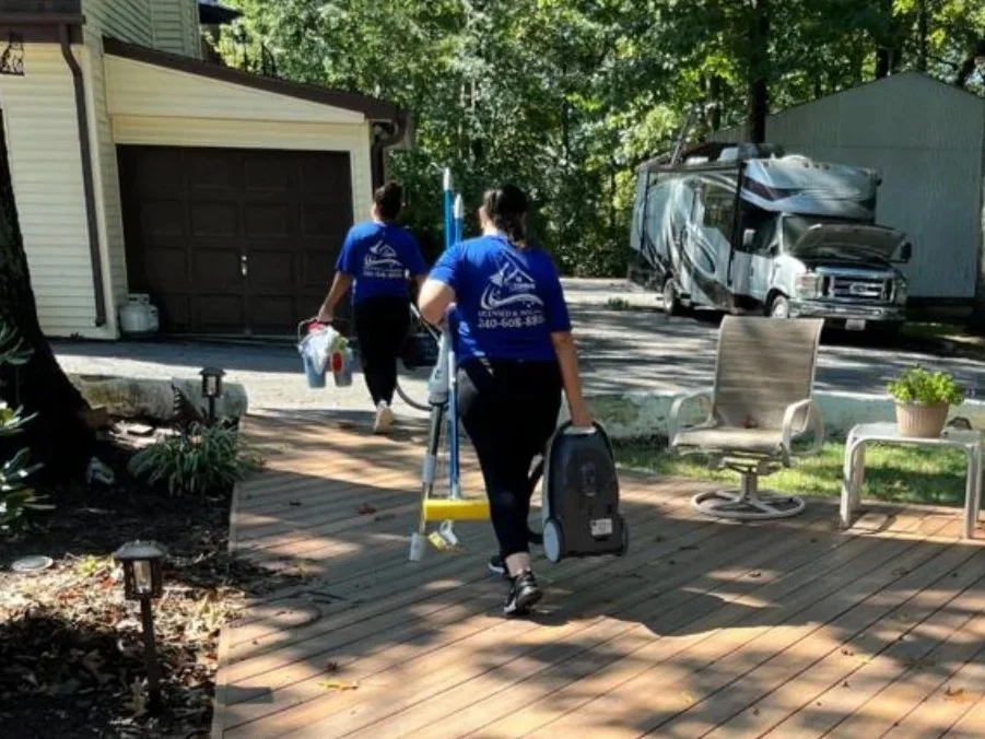 Two people wearing matching blue shirts walking away from the camera on a wooden deck, carrying cleaning supplies and equipment, with a suburban house, a garage, a recreational vehicle, garden furniture, and trees in the background.