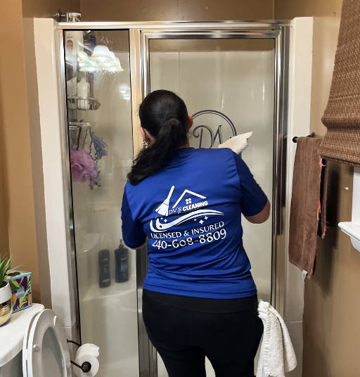 A woman in a blue shirt cleaning the inside of a glass shower door in a bathroom.