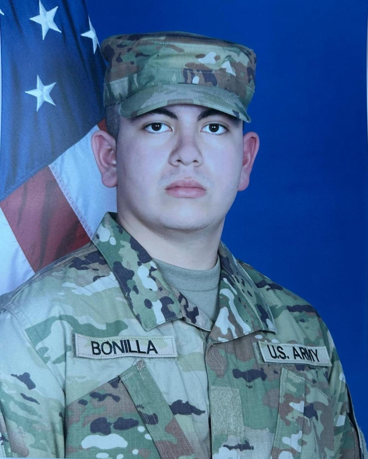 A young U.S. Army soldier in camouflage uniform and cap, standing in front of an American flag. The uniform has name tag 'BONILLA' and 'U.S. Army' patches.