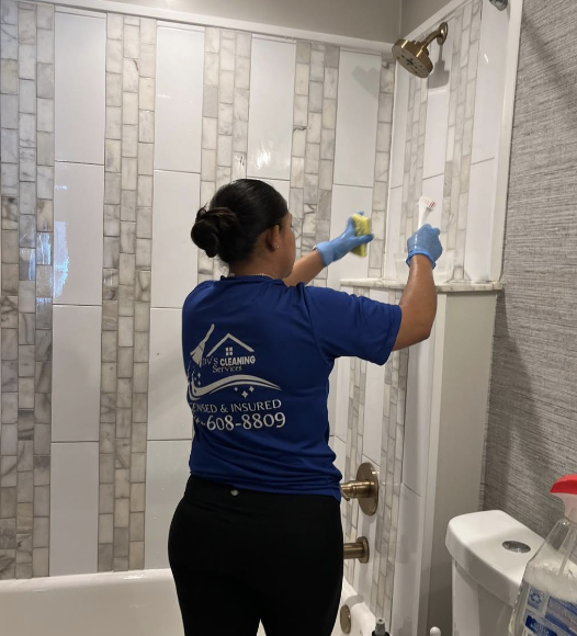 A woman cleaning a bathroom shower wall with a sponge, wearing a blue shirt and gloves.