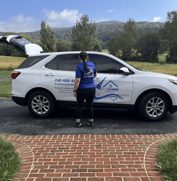 A woman standing next to a white SUV with the hatchback open, in a rural area with trees and hills in the background. The vehicle is branded with a cleaning service logo.