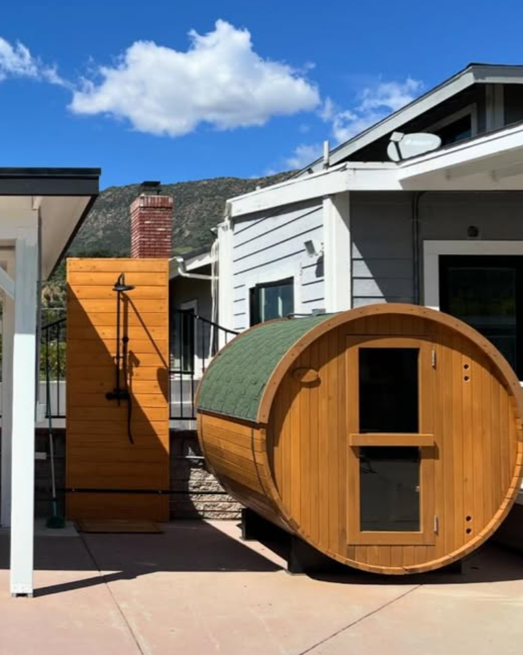 Custom outdoor barrel sauna built by Repairs y More featuring beautiful cedar wood construction, a green shingled roof, and an adjacent outdoor shower station in a Los Angeles area backyard.