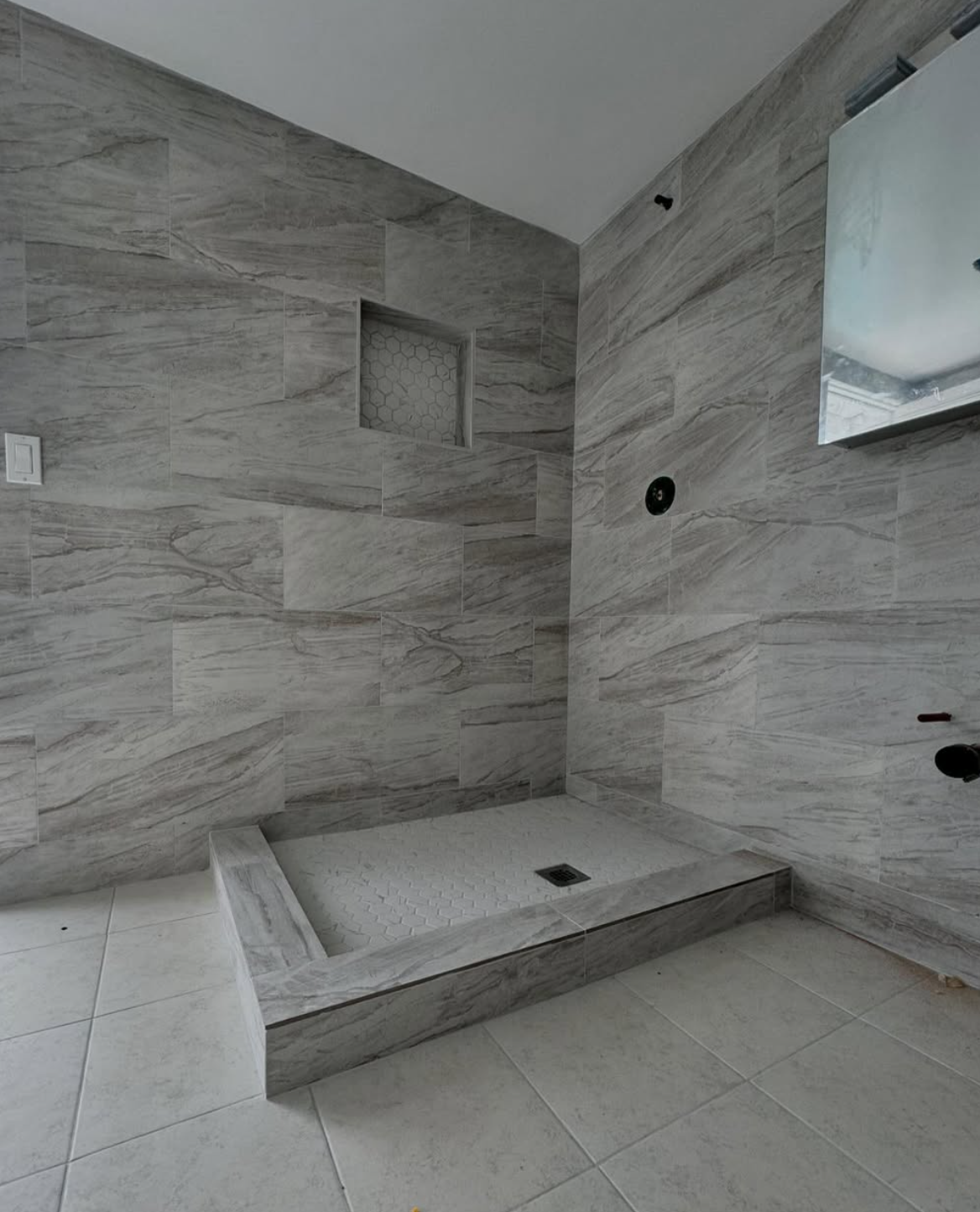 Modern walk-in shower renovation by Repairs y More featuring large-format grey marble-look tiles, hexagon tile accent niche, and clean white herringbone floor tile in a Los Angeles area bathroom.