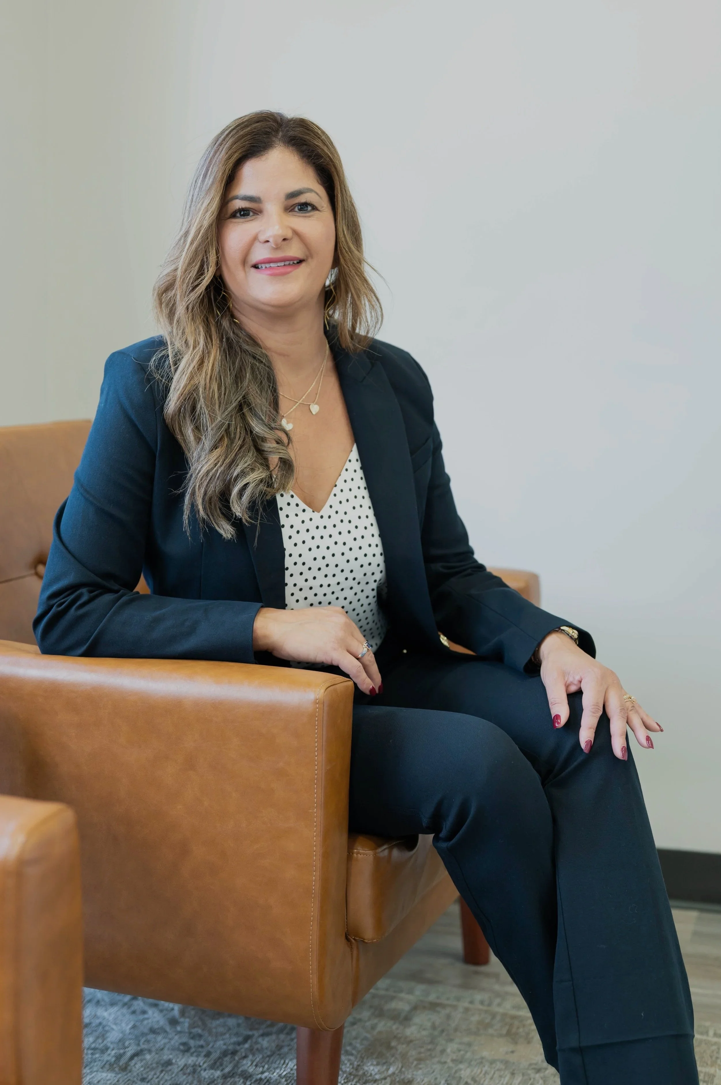 A woman with wavy brown hair, dressed in a navy blazer and polka dot blouse, sitting on a brown leather chair in a room with a plain, off-white wall.