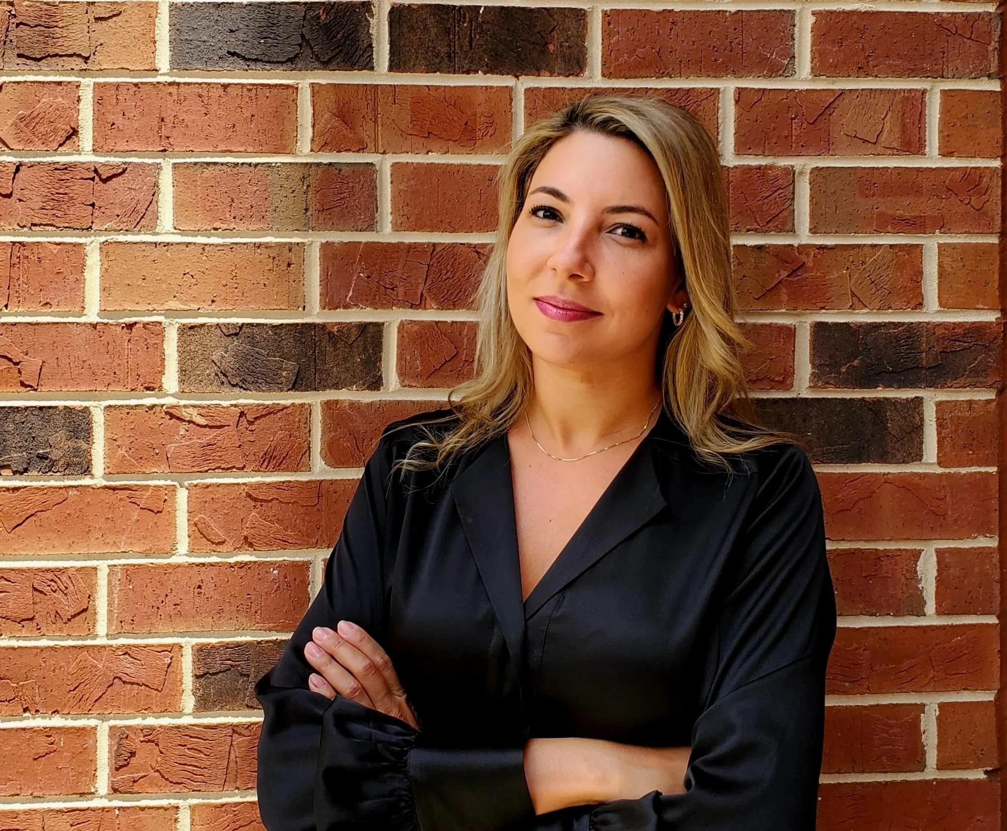 A woman with blonde hair, wearing earrings, a black top, and a necklace, standing in front of a brick wall with her arms crossed.
