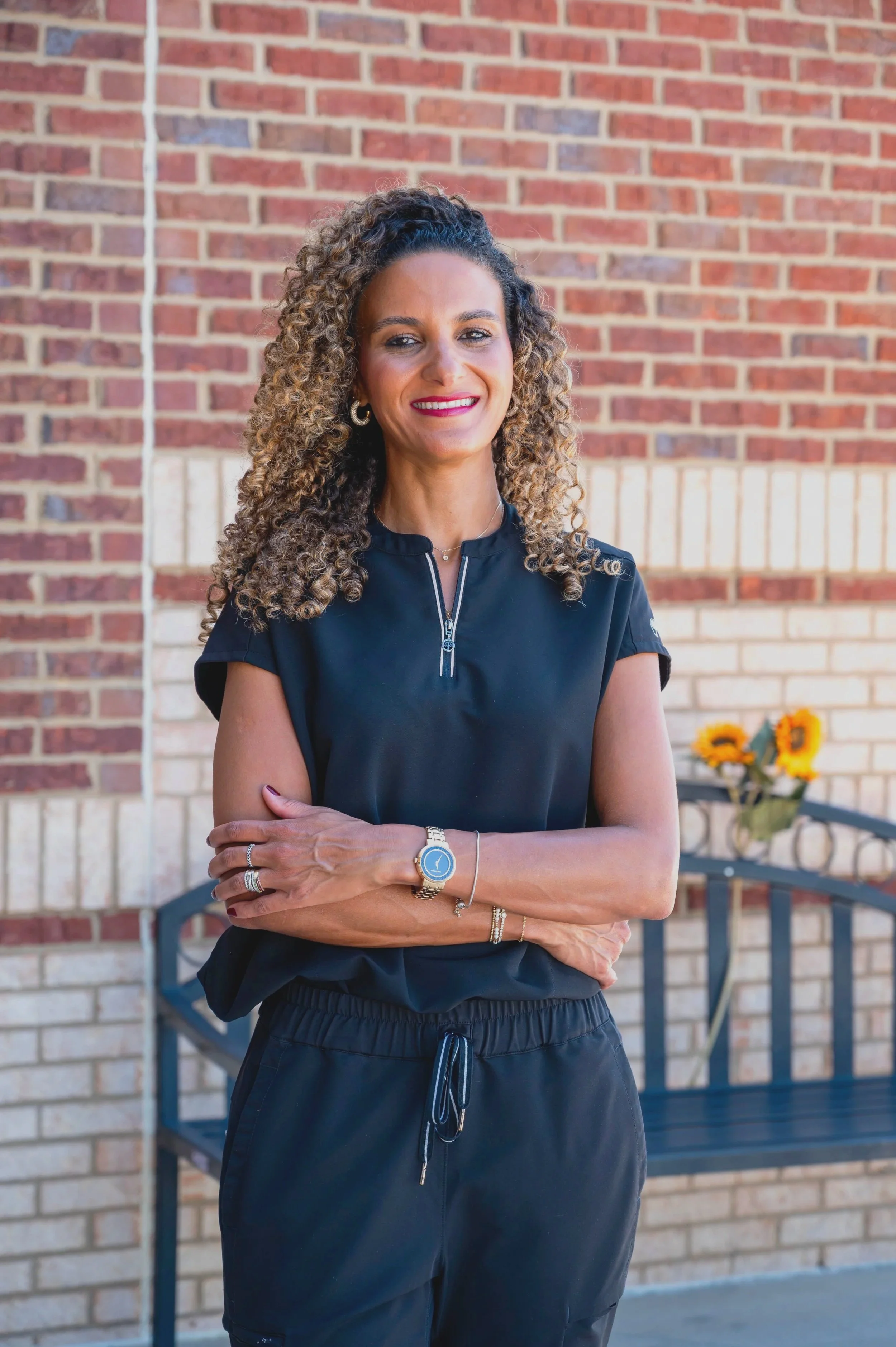 A woman with curly hair, wearing a black shirt with a zipper, gold jewelry, and a watch, standing outdoors with her arms crossed, smiling in front of a brick wall and a bench with sunflowers in the background.