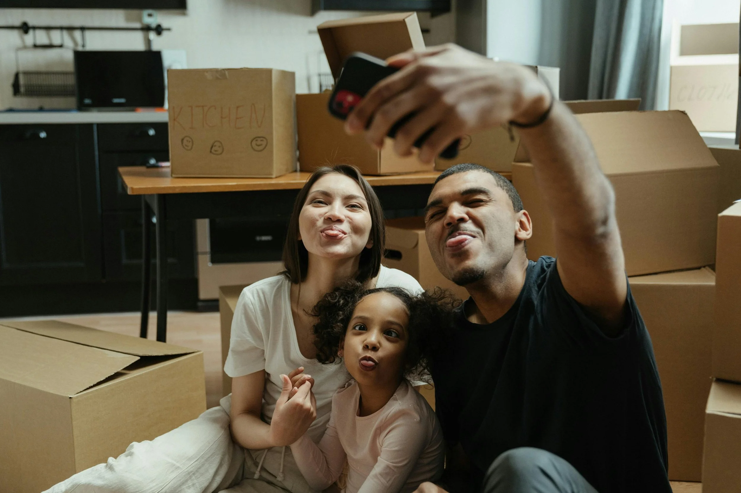 A family of three taking a selfie inside a room with moving boxes around them, making funny faces.