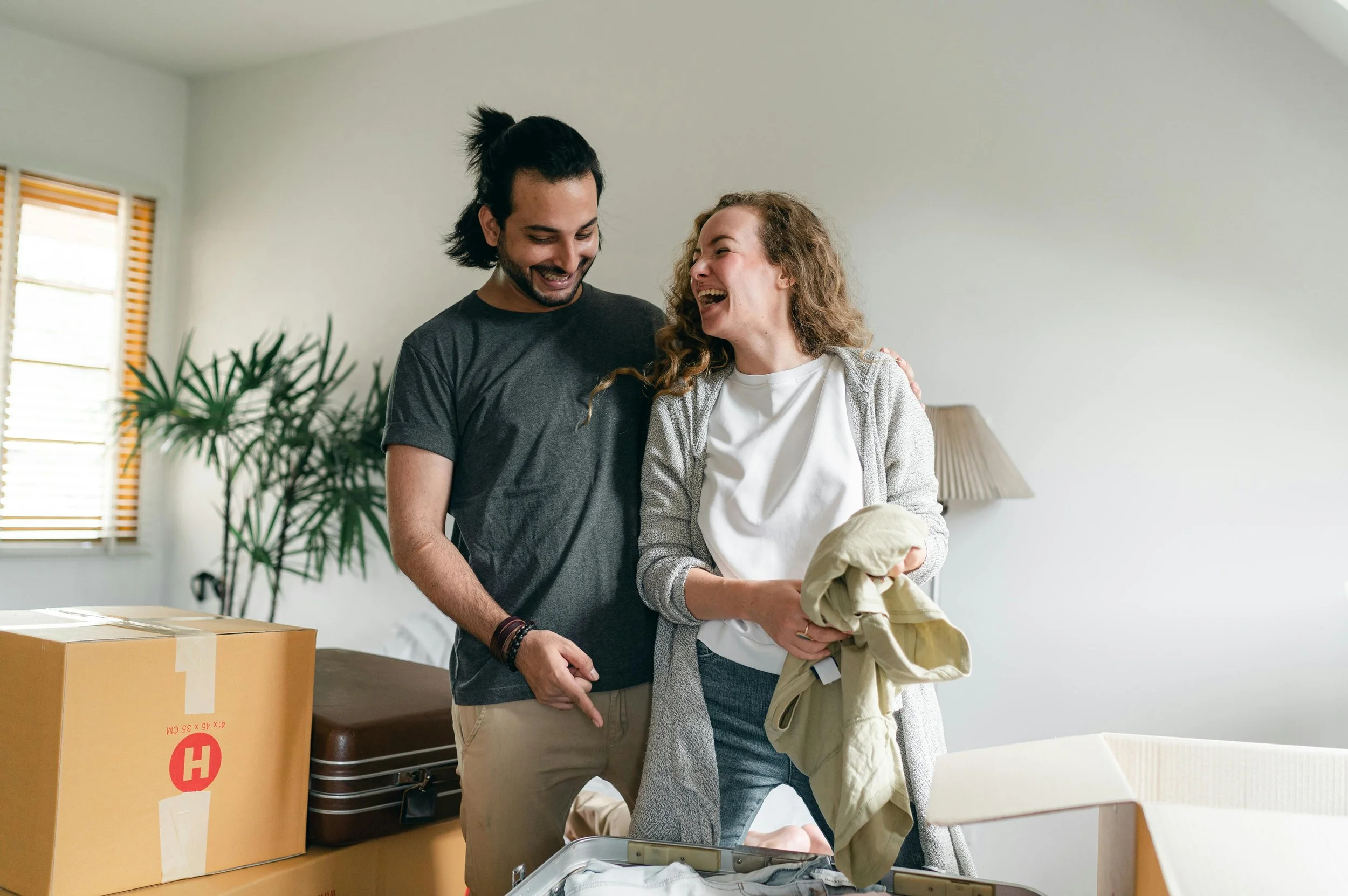 A smiling couple packing clothes and standing next to a cardboard box in a bright living room with a plant and a window.