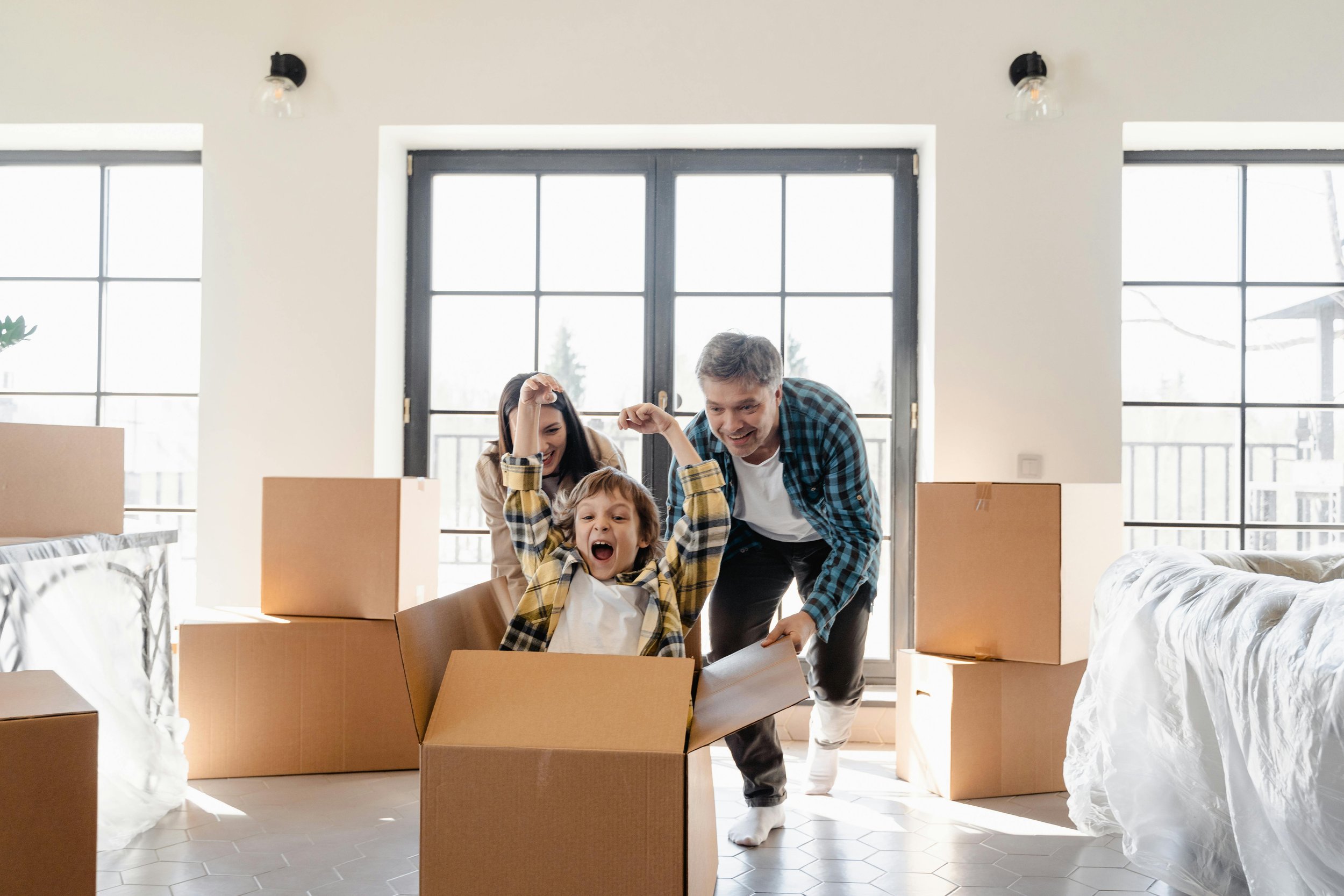 A happy family moving into a new home, with a boy celebrating in a cardboard box and parents smiling around him, amid unpacked boxes and large windows.