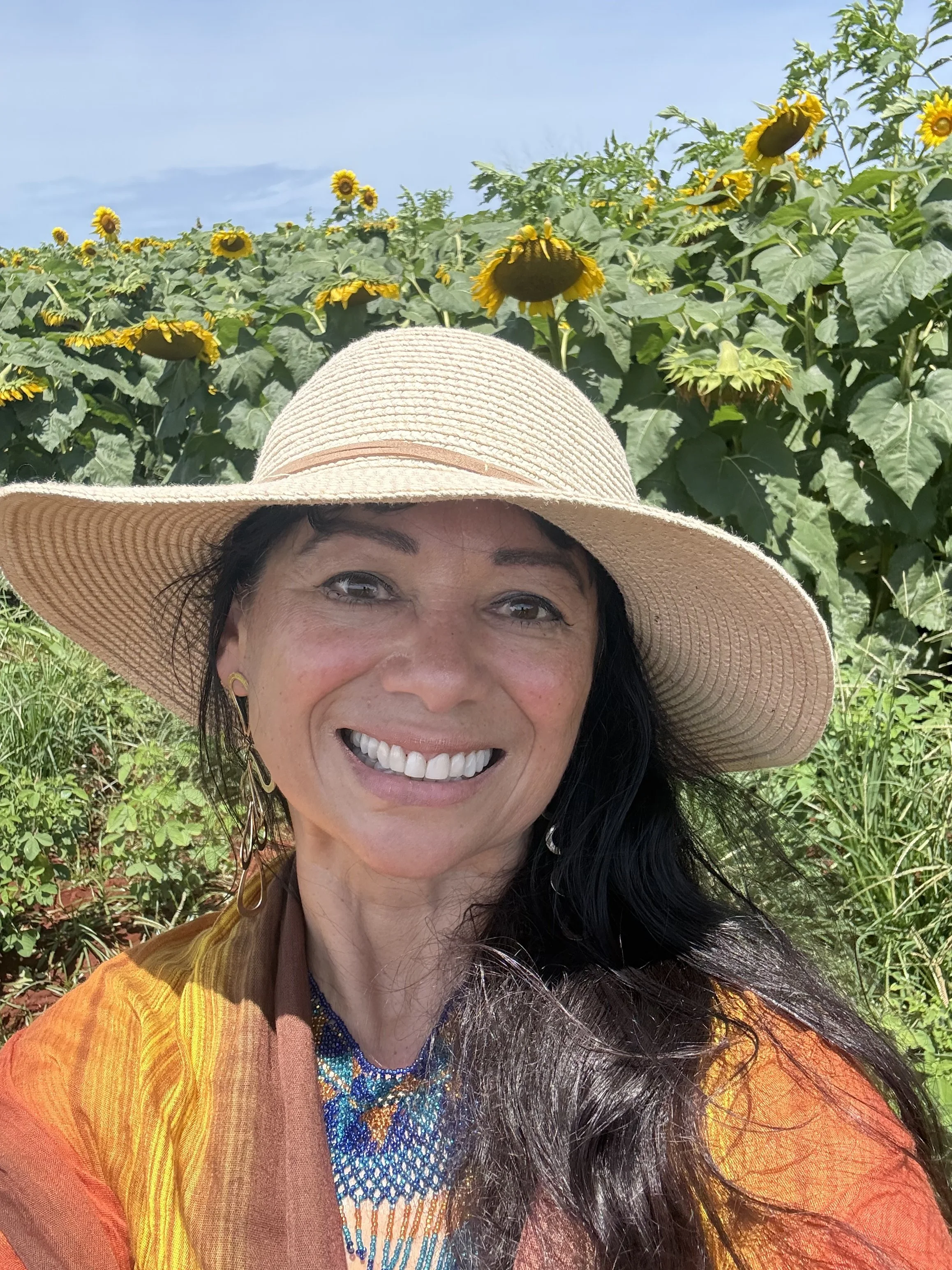 A woman wearing a large sunhat and colorful, layered clothing taking a selfie in front of a sunflower field under a blue sky.