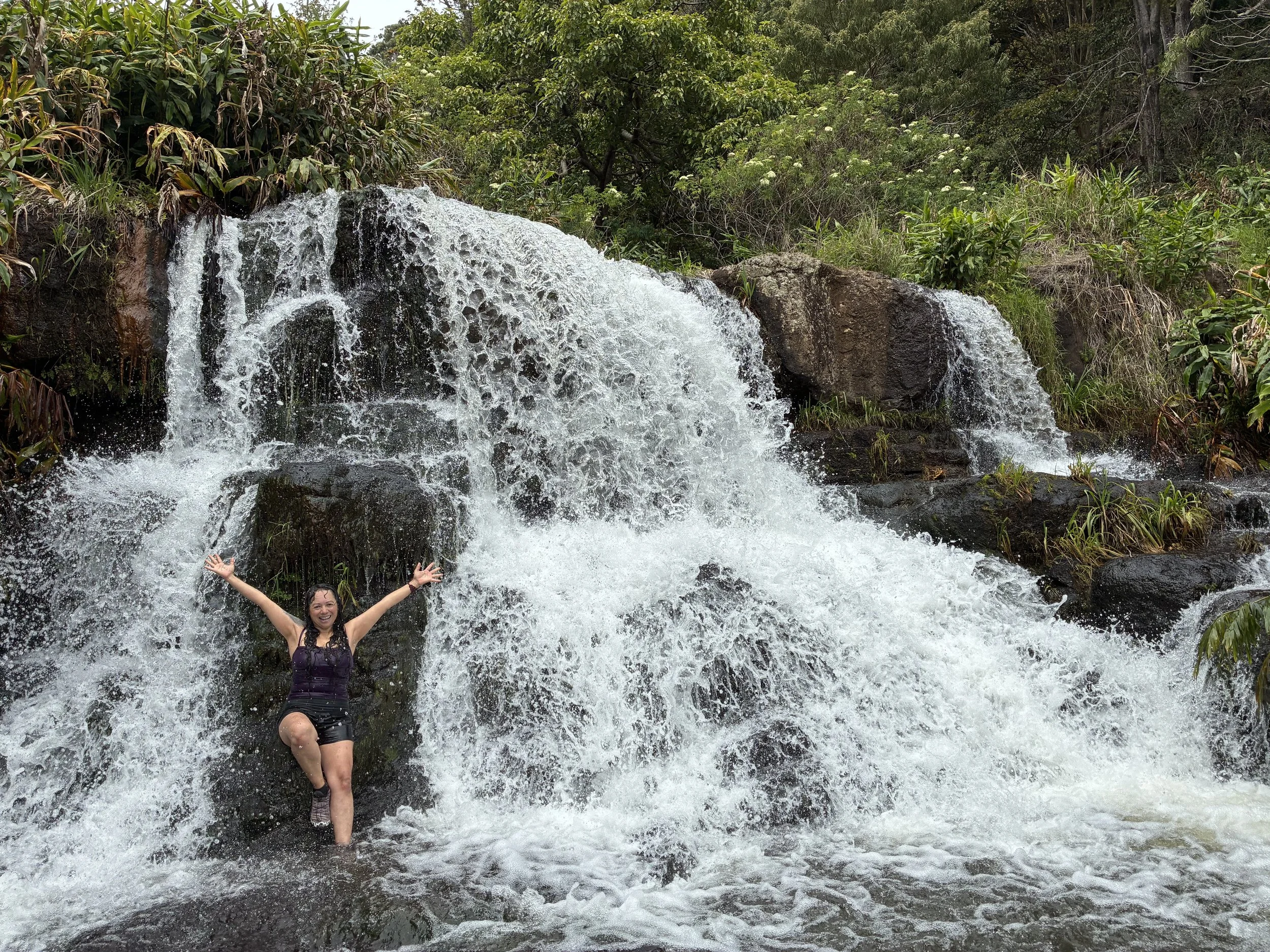 A woman is standing with arms raised in front of a waterfall in a lush green forest. She appears to be happy and enjoying the outdoor scenery.