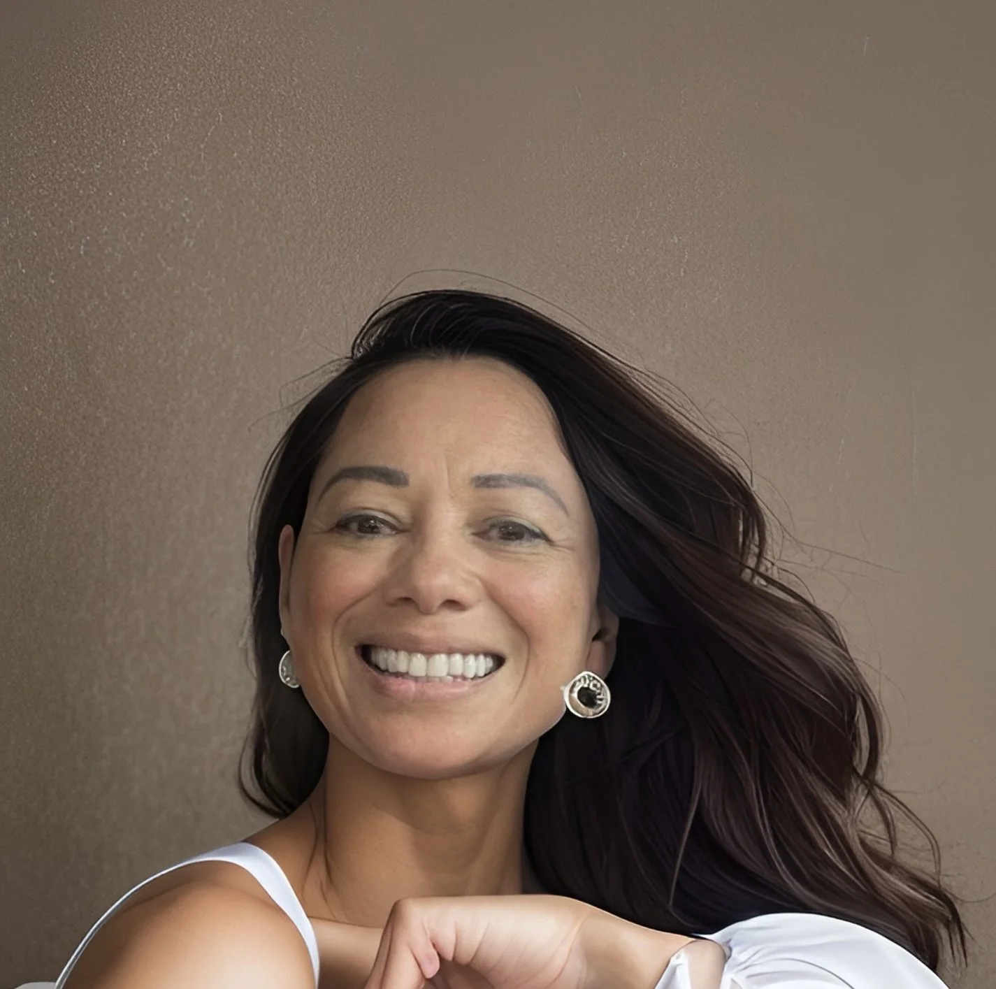 A smiling woman with long brown hair, wearing earrings and a white top, standing against a beige wall.