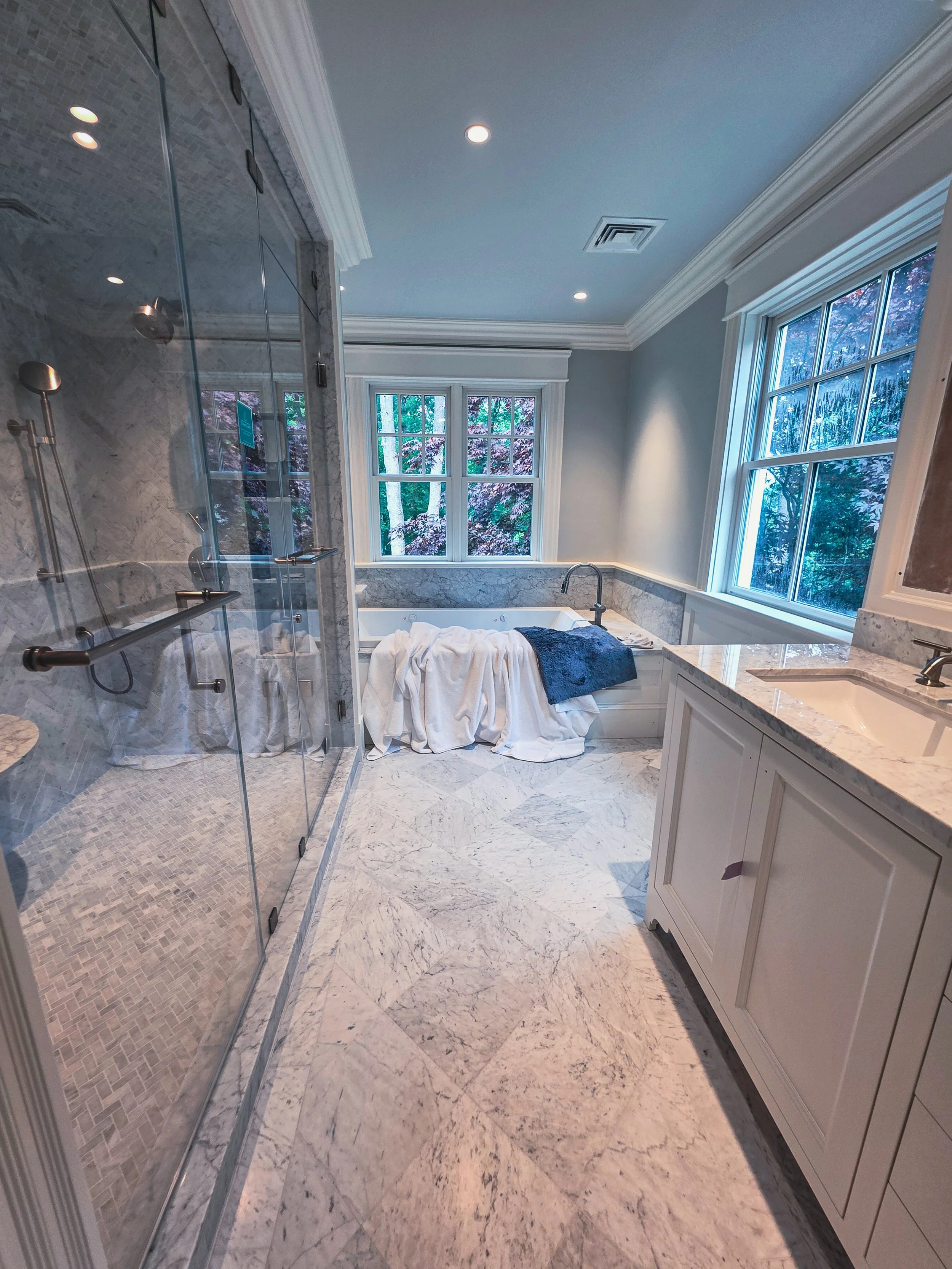 A bathroom with a glass-enclosed shower on the left, marble floors, and a built-in bathtub under large windows showing trees outside. White cabinetry and granite countertops are on the right.