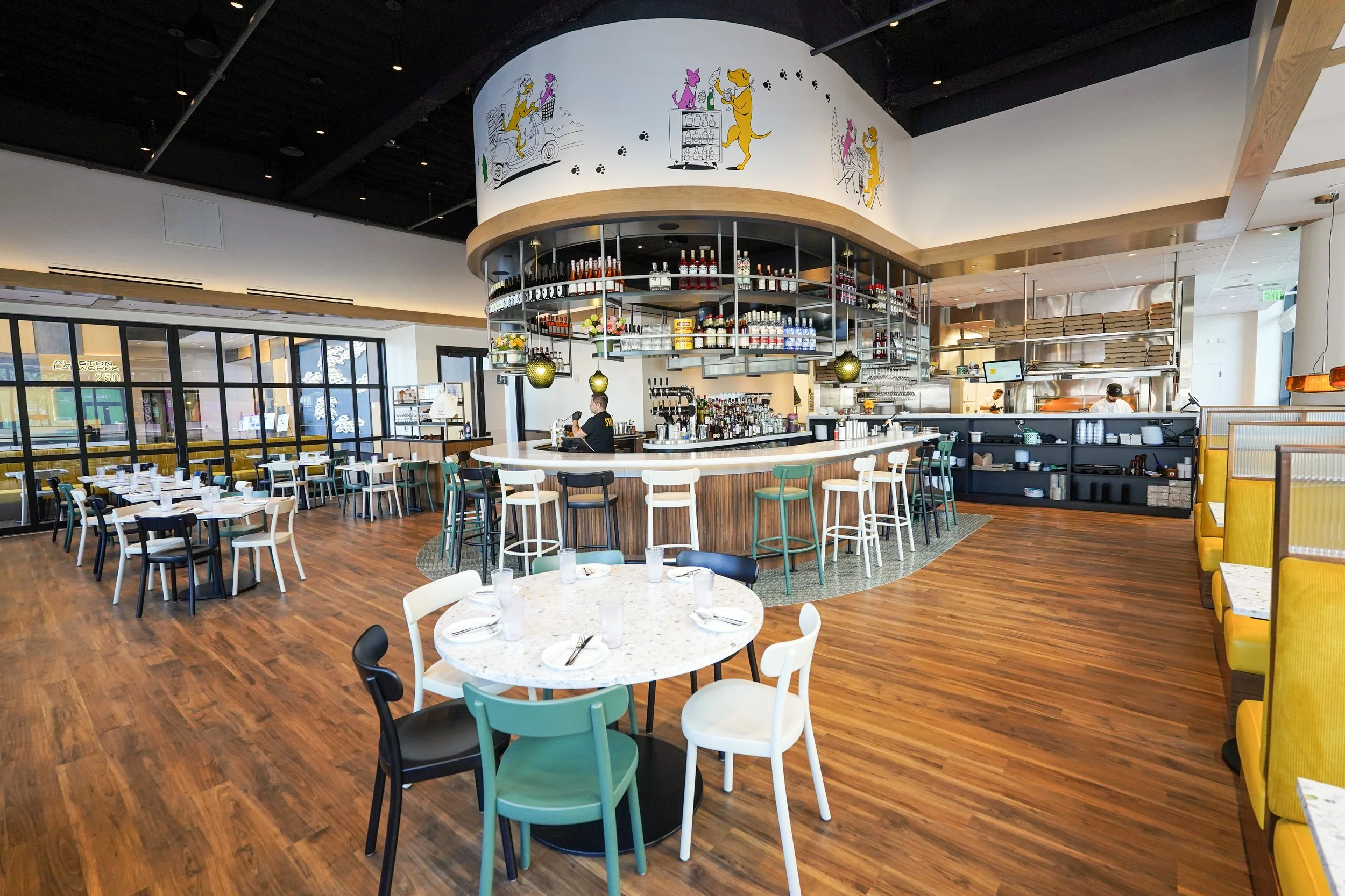 Modern restaurant interior with colorful chairs and round tables, a bar area with bottles and glasses, and a mural of cartoon dogs on the curved ceiling