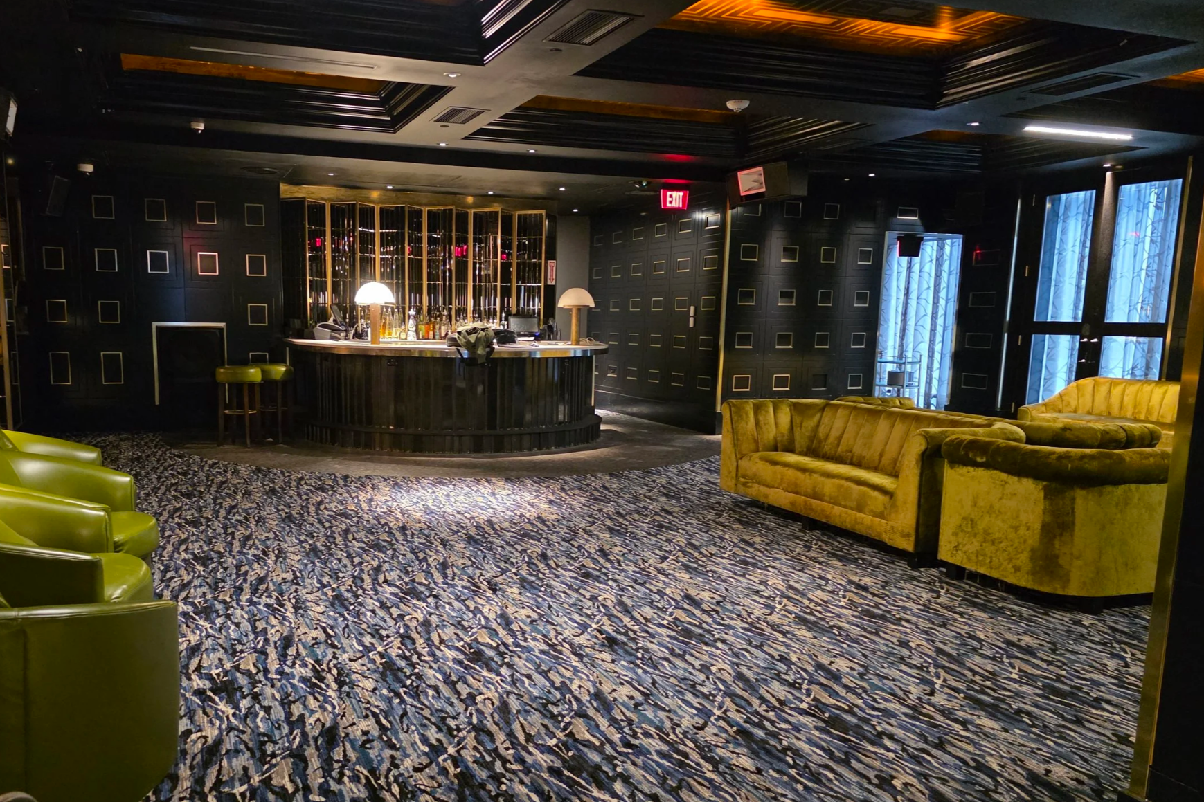 An elegant dimly lit lounge area with a black bar in the center, green and yellow velvet sofas, a patterned carpet, and large windows with blue curtains.