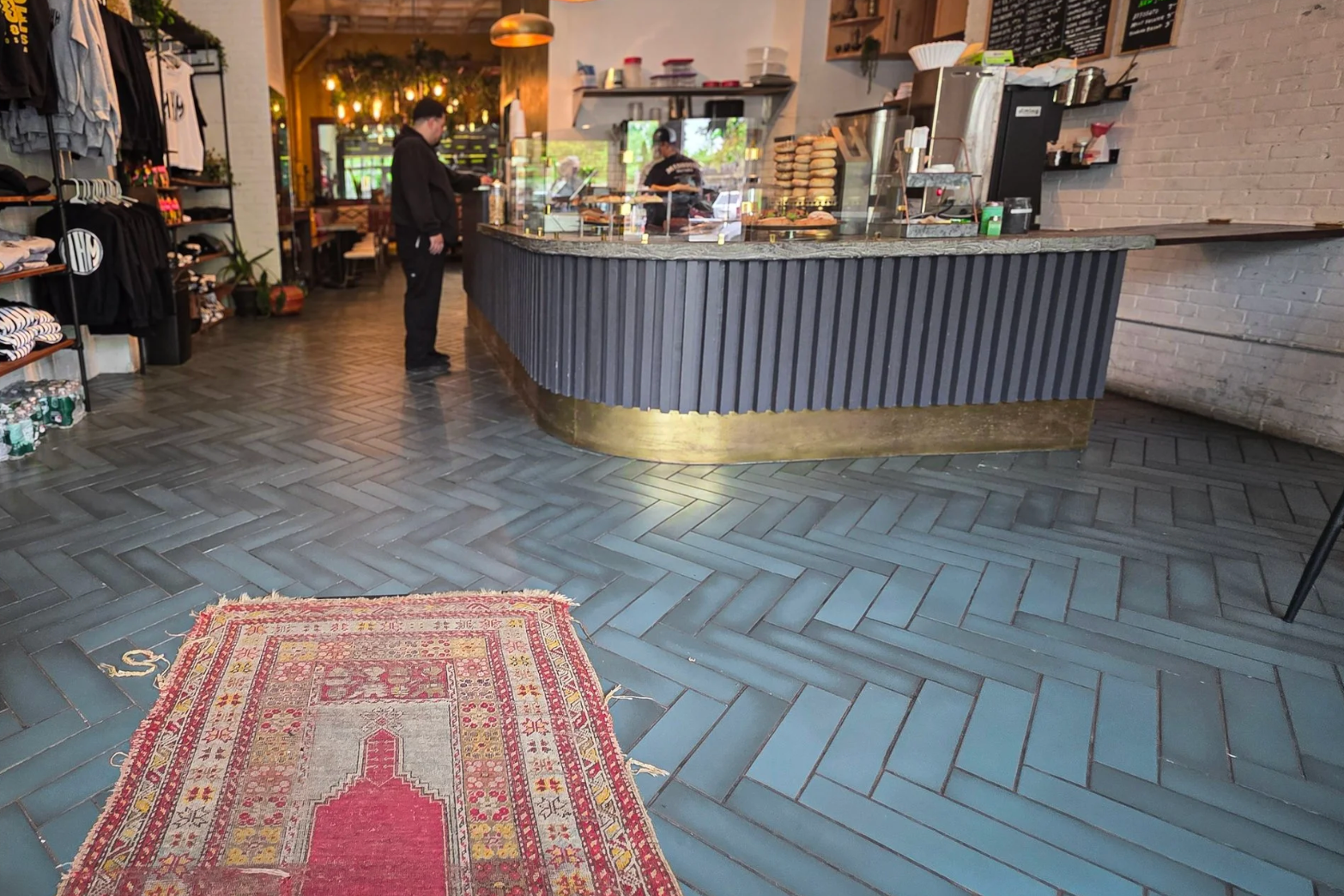 Interior of a modern cafe or coffee shop with a curved counter, two employees working behind it, wooden shelves with baked goods, and clothing and merchandise on display on the left side.