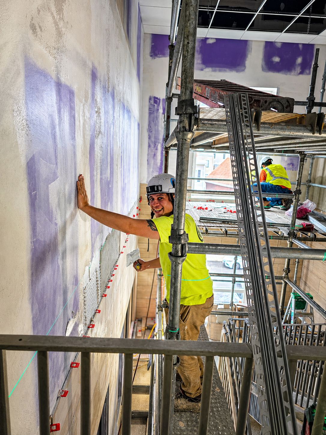 Construction worker smiling and working on a wall with purple markings, standing on scaffolding inside a building under renovation.