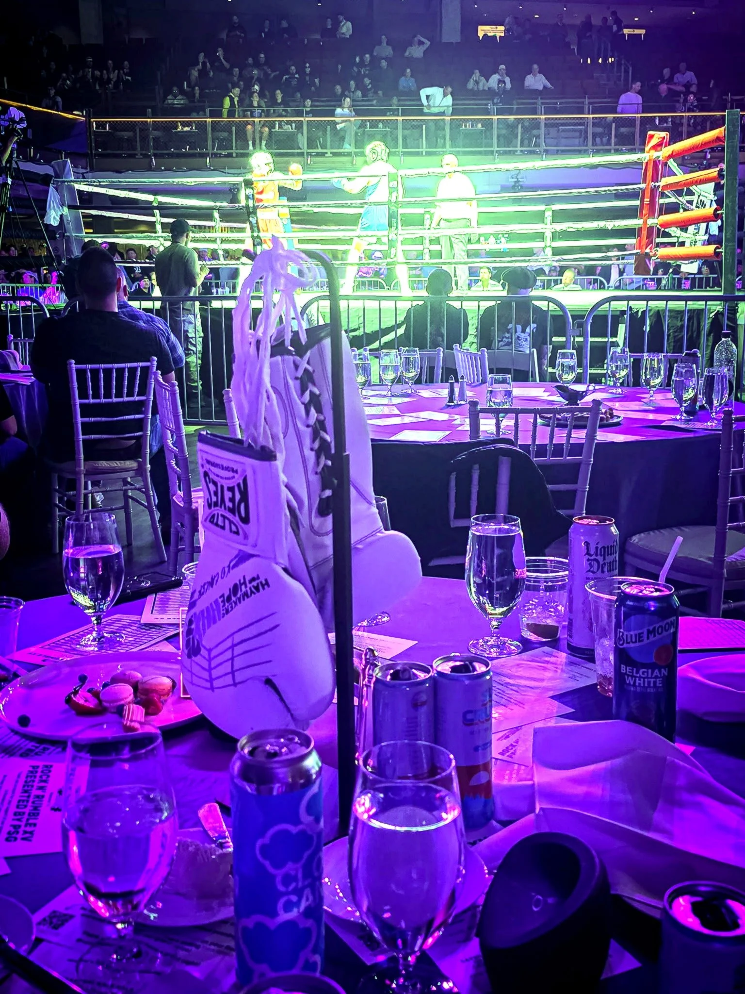 Boxing match in a large arena with an audience, view from a dining table with glasses, drinks, and snacks, illuminated by purple and green lights.