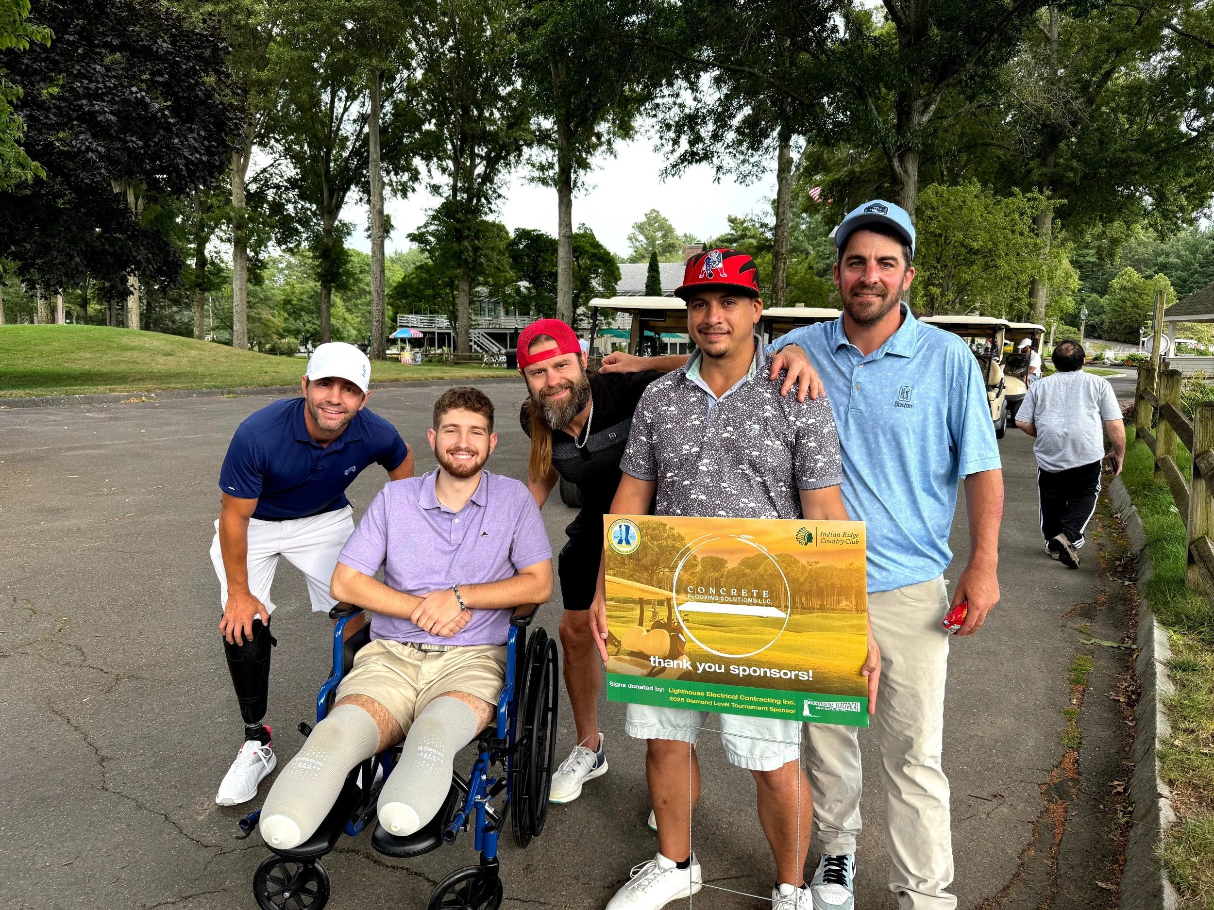 A group of five men, including one in a wheelchair, standing outdoors in a park, holding a sign that reads "thank you sponsors" at a golf event, with trees and golf carts in the background.