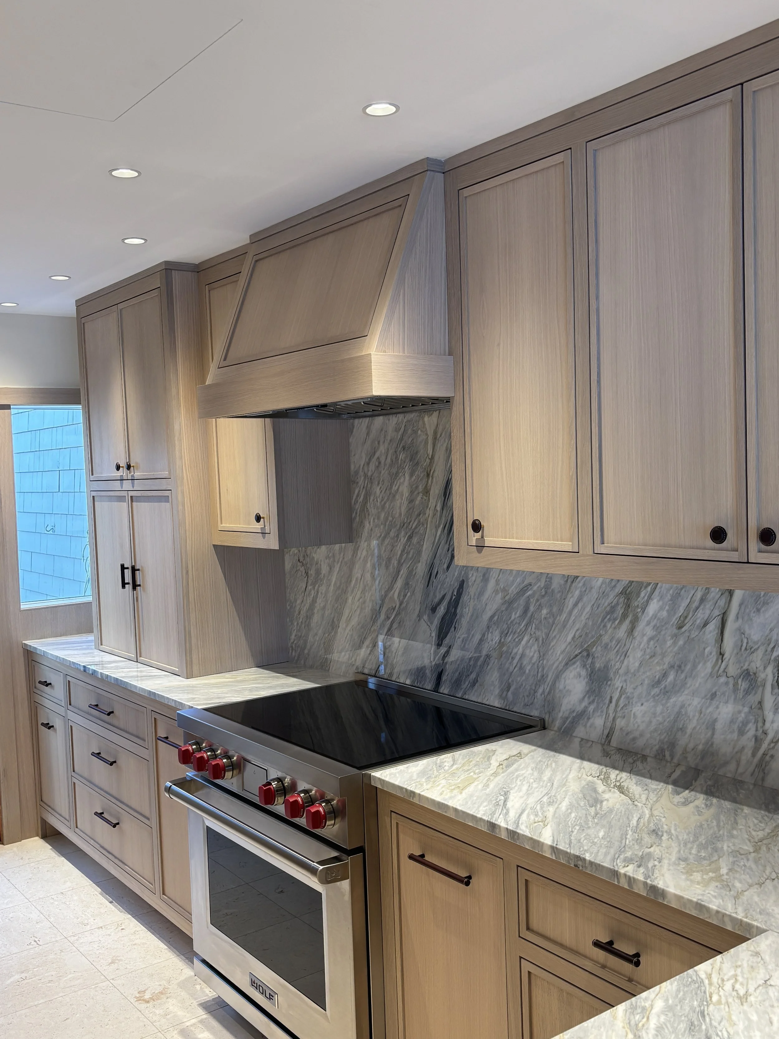 View of a modern kitchen with light wood cabinets, marble countertops, a black stovetop, and a stainless steel oven, with recessed lighting in the ceiling.