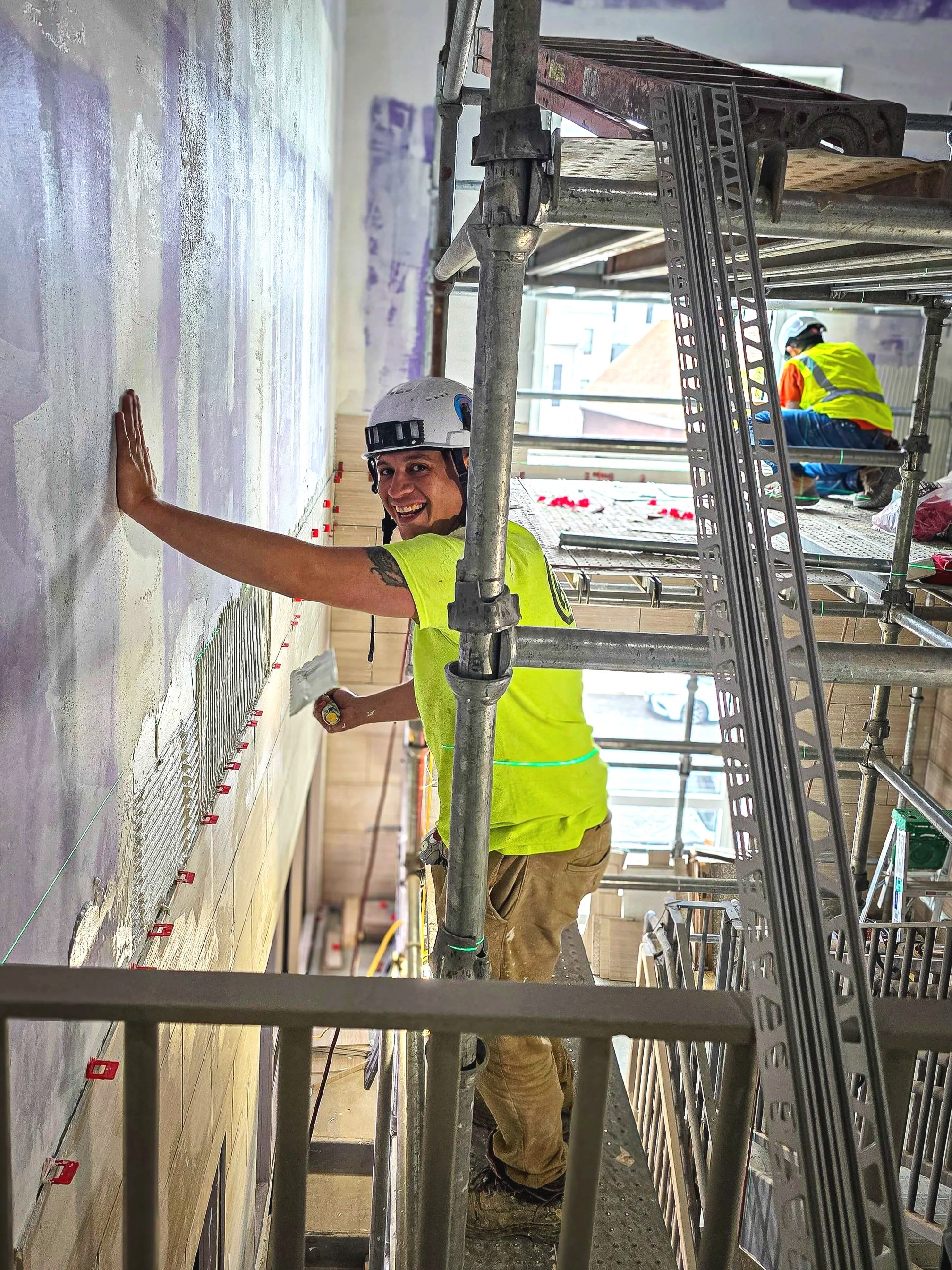 A construction worker wearing a white safety helmet and a bright yellow shirt working on a wall inside a building. The worker is smiling and looking at the camera. There is scaffolding around him and another worker in the background wearing a safety vest and helmet.